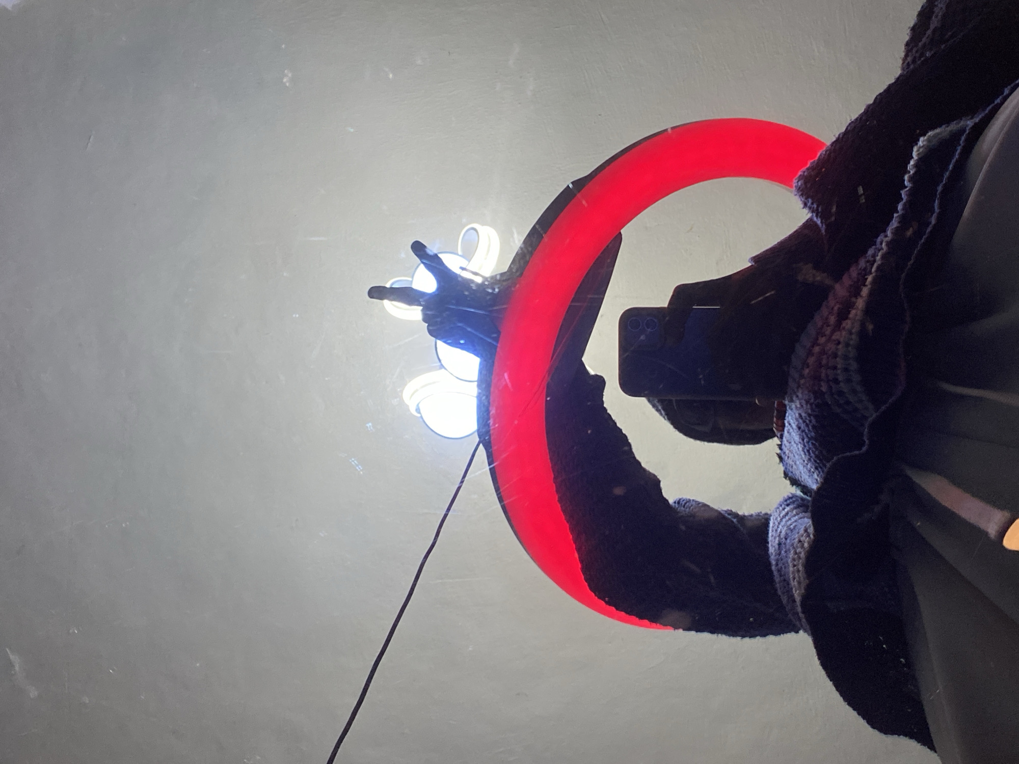 This is a low-angle, waist-up, waist-level shot of a person holding a smartphone in their right hand, reflected in a large, circular, red neon light sign. The sign is mounted on a white, textured wall, possibly in an establishment in Abakaliki, Nigeria. The person is wearing dark clothing and a knitted scarf or shawl in shades of blue and purple. Their left arm is also visible, covered by a dark, possibly knitted sleeve. The smartphone is positioned to capture the reflection of the neon sign and the person's face, which is partially obscured. The sign itself is bright red and emits a strong glow, illuminating the surrounding area with a warm light. Several smaller, white lights are visible above the red sign, likely part of the same installation. A thin black wire stretches diagonally across the upper left portion of the image. The overall impression is of a candid, possibly artistic, self-portrait or a moment captured while exploring a brightly lit urban space.