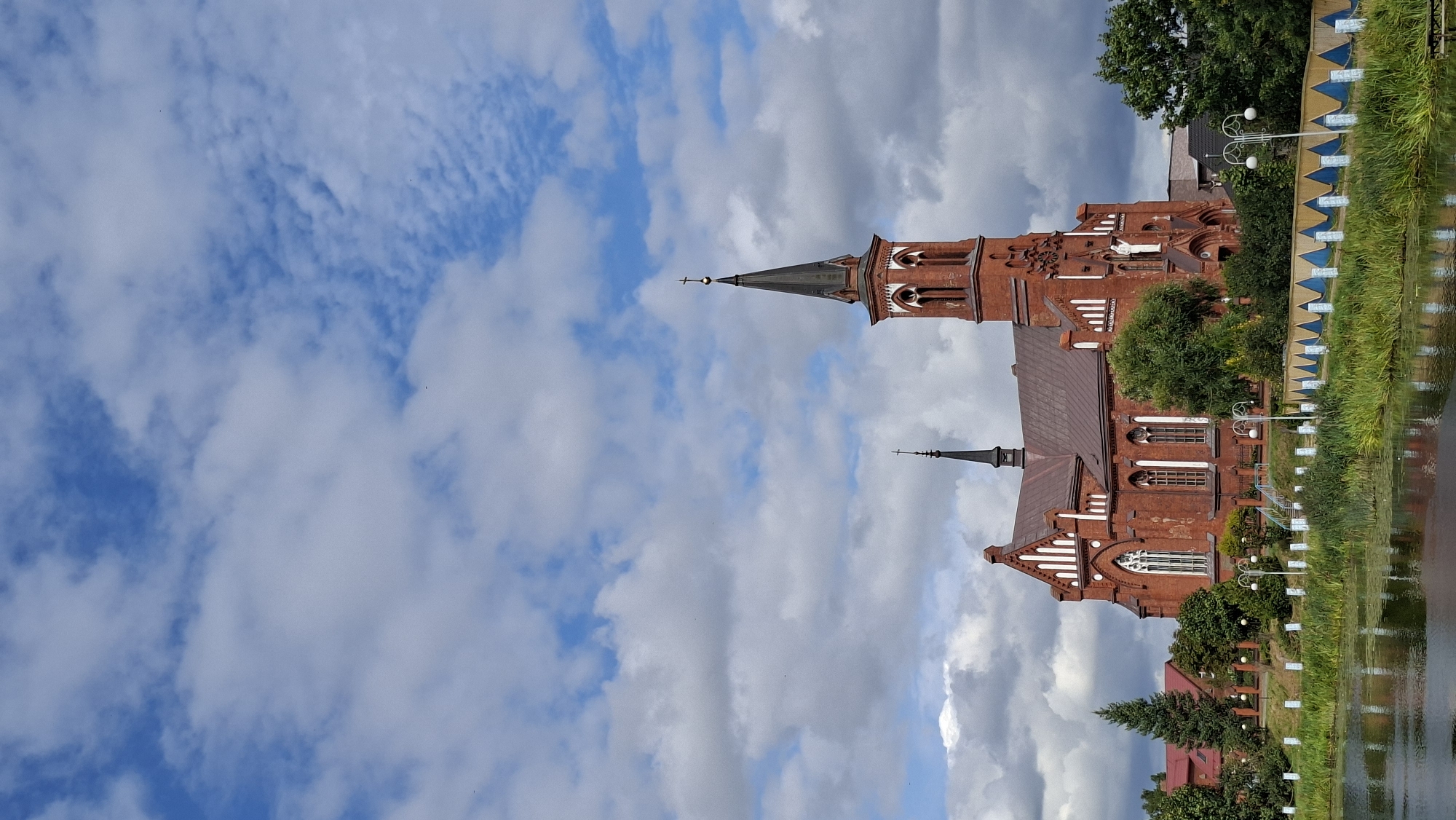 The image depicts the Church of the Transfiguration of the Lord in Pastavy, Belarus. The brick church, characterized by its neo-Gothic architectural style, is the central focus. It features a tall, slender spire topped with a cross, and a prominent main tower. The church is situated on the bank of a body of water, likely a lake or river, with green reeds and vegetation growing along the shoreline. A decorative fence with blue and yellow accents runs along the water's edge. In the background, there are trees and some buildings. The sky is partially cloudy, with patches of blue sky visible between the white and gray clouds. The overall scene is that of a historical landmark in a tranquil, natural setting under a dynamic sky.