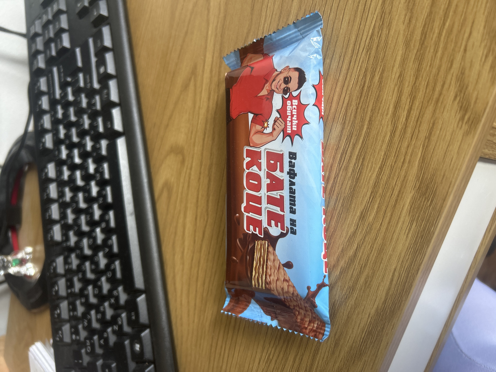 The image shows a partially opened "BATE KOCE" wafer bar lying on a light brown wooden surface. The bar has a light blue and brown wrapper with a cartoon image of a muscular man on the front. The brand name is printed in large red letters, and some Cyrillic text is also visible. The wafer bar is partially obscured by a black keyboard to the left of the frame. The scene appears to be indoors, potentially on a desk or table. No specific details indicate a location in Burgas, Bulgaria, or the involvement of any people beyond the cartoon on the packaging.