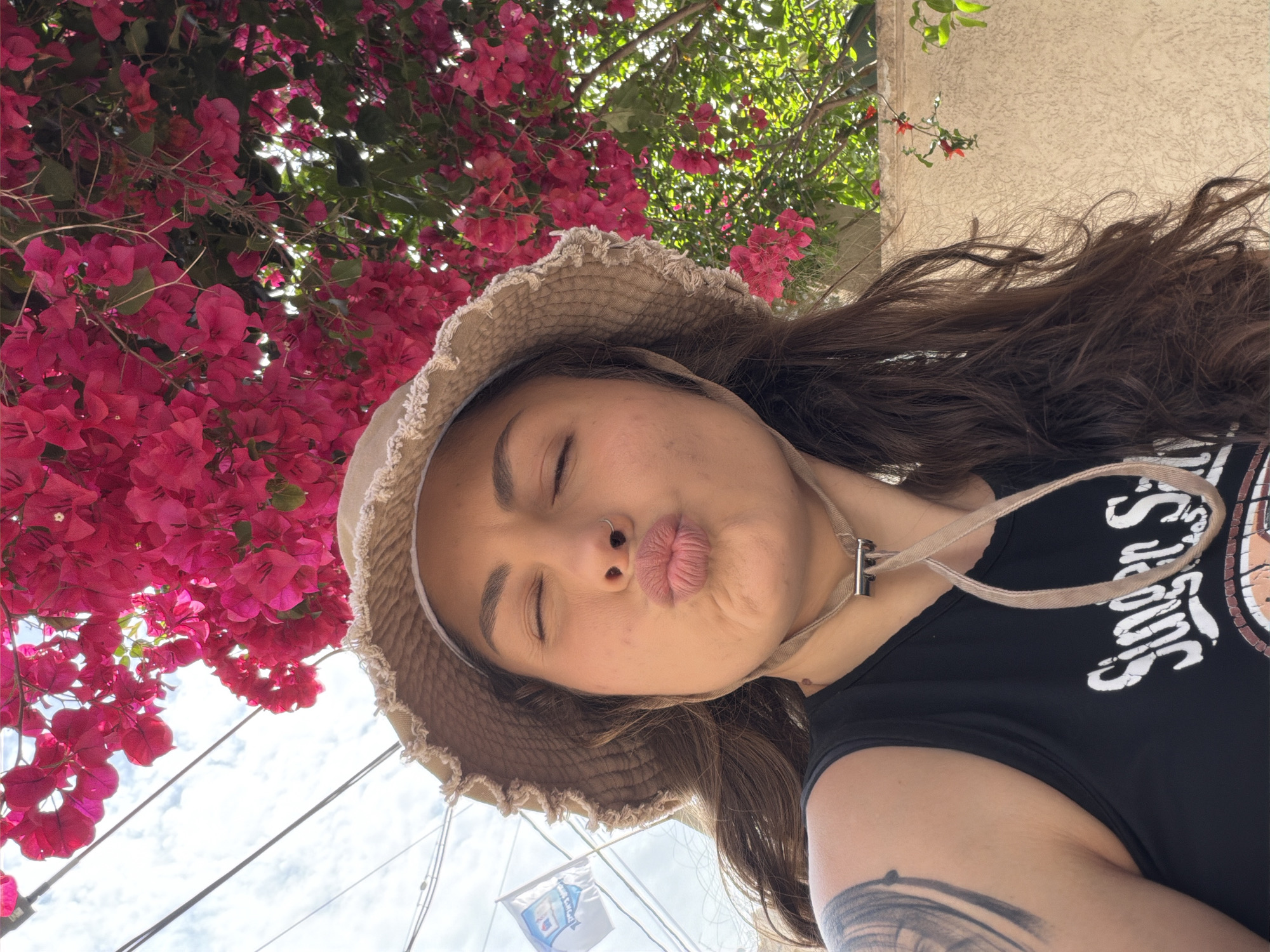 A selfie shows a person with closed eyes, a nose ring, and pursed lips, wearing a beige hat with a drawstring around their neck and a black t-shirt with a white graphic. The person's brown hair is partially visible under the hat. The background includes vibrant pink bougainvillea flowers and green foliage. A blue and white flag is visible, with power lines running in front of a cloudy sky. The location is Kiryat Bialik, Israel.