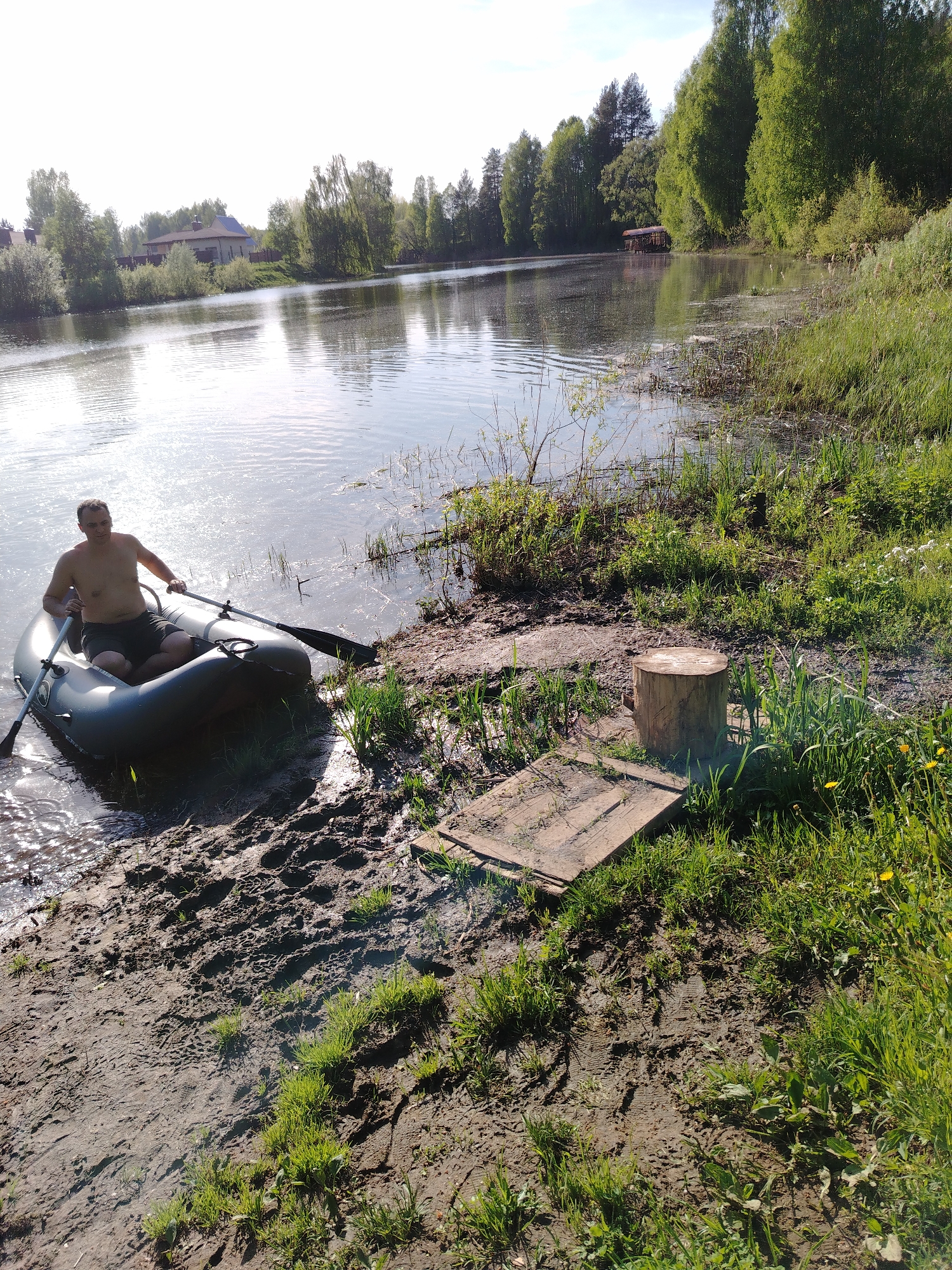 A man with a bare chest is in a gray inflatable boat near the muddy shore of a body of water. He holds an oar in each hand, presumably to propel the boat. To the right of the boat, on the shore, is a wooden platform and a tree stump. Green grass and vegetation cover the bank, blending with the water's edge. In the background, trees line the opposite shore, with visible buildings and a structure on the water. The photo was taken in the city of Stroitel', Russia.