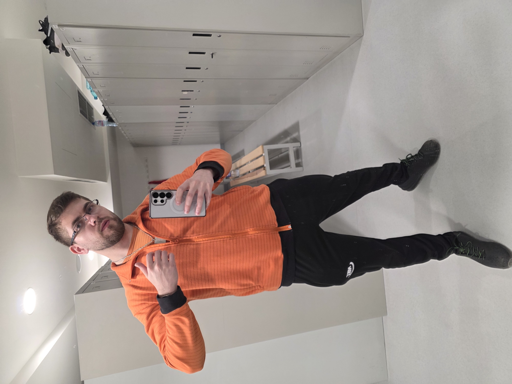 A man is taking a selfie in a locker room. The room has rows of metal lockers. The man is wearing glasses, an orange jacket, and black Nike sweatpants. He is holding a smartphone. The locker room appears to be sparsely furnished. It is impossible to verify the location as Plovdiv, Bulgaria from the provided image.