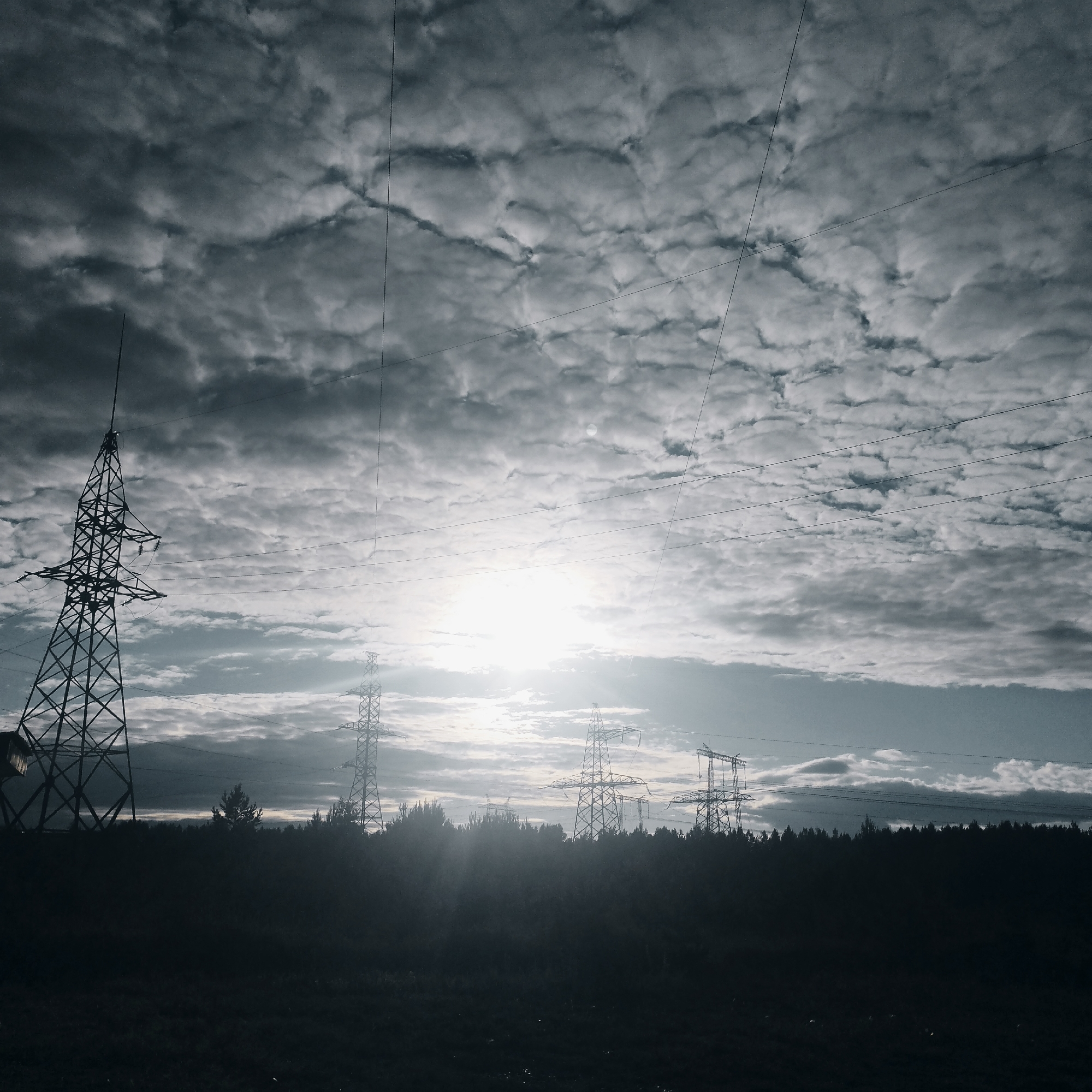 The image depicts a rural landscape with a dramatic sky filled with textured clouds. In the foreground, silhouetted against the bright sky, are several tall metal electricity pylons and a line of dark trees. The pylons are lattice-like structures, characteristic of high-voltage power lines. The sun is visible, partially obscured by clouds, casting rays of light through the sky. The overall impression is one of an industrial element integrated into a natural, somewhat moody environment. The location is specified as the village of Pokosnoye, Russia. There are no people visible in the image.
