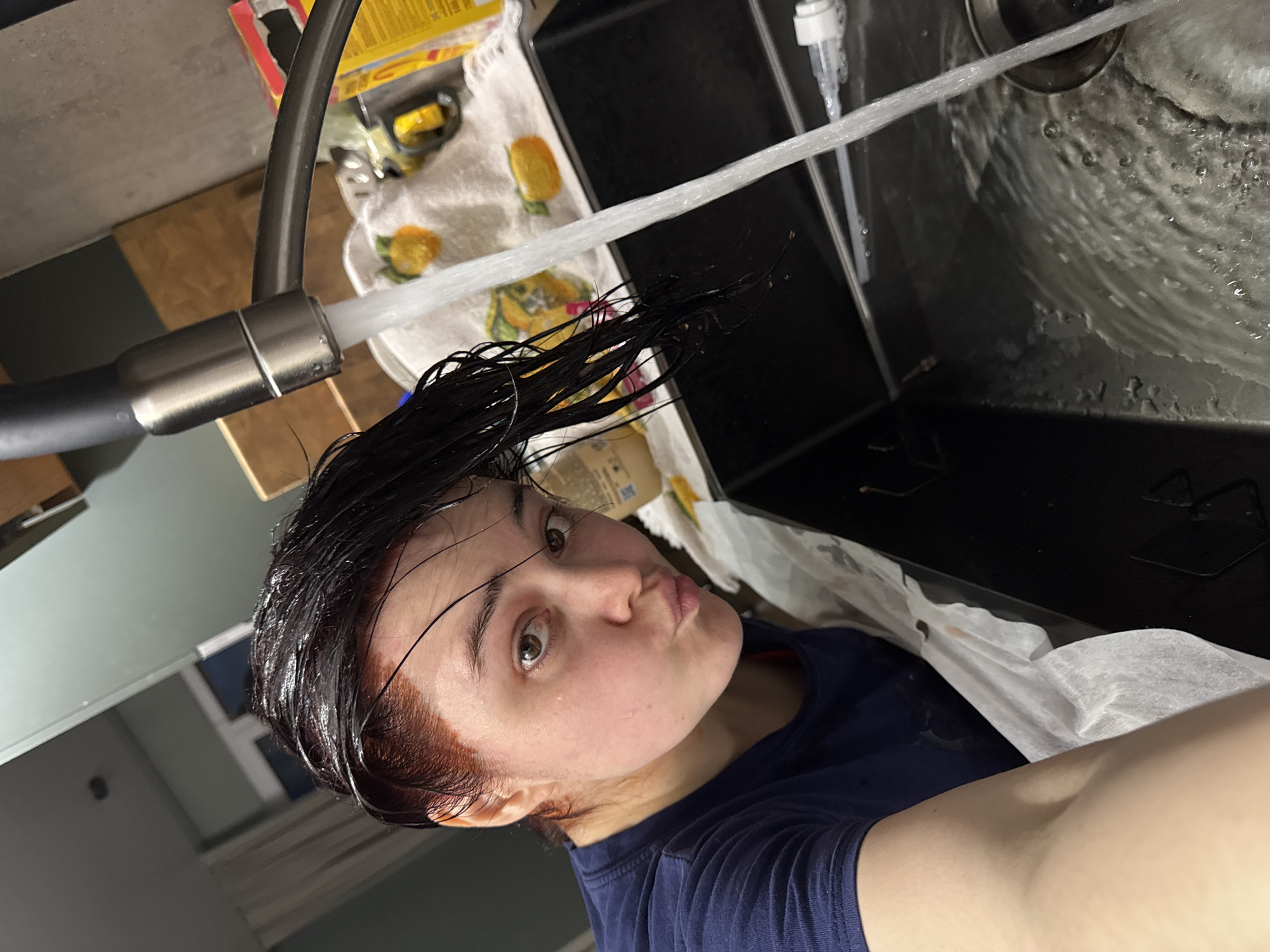 This vertical image depicts a young person rinsing their dark, wet hair in a kitchen sink, likely at home in Izhevsk, Russia. The primary subject is a person, possibly female, with a "duck face" expression, looking directly at the camera. Her eyes are wide open, and some strands of her dark, wet hair are visible on her forehead. A noticeable reddish tint around her hairline and scalp suggests she is rinsing hair dye. She is wearing a dark blue t-shirt.
The person is leaning over a dark-colored kitchen sink where a strong stream of water flows from a modern kitchen faucet with a pull-out spray head. The water creates splashes and ripples in the sink. To the right of the sink, a white pump dispenser bottle, likely containing soap or shampoo, is visible. In the background, a white, possibly patterned towel or cloth rests on the counter, adorned with a few yellow items. Further back, blurred containers, including a yellow box with unreadable text, are present. The upper left portion of the image shows a reflection, possibly of the ceiling or other parts of the room. The scene captures a candid, everyday moment during a hair care routine.
