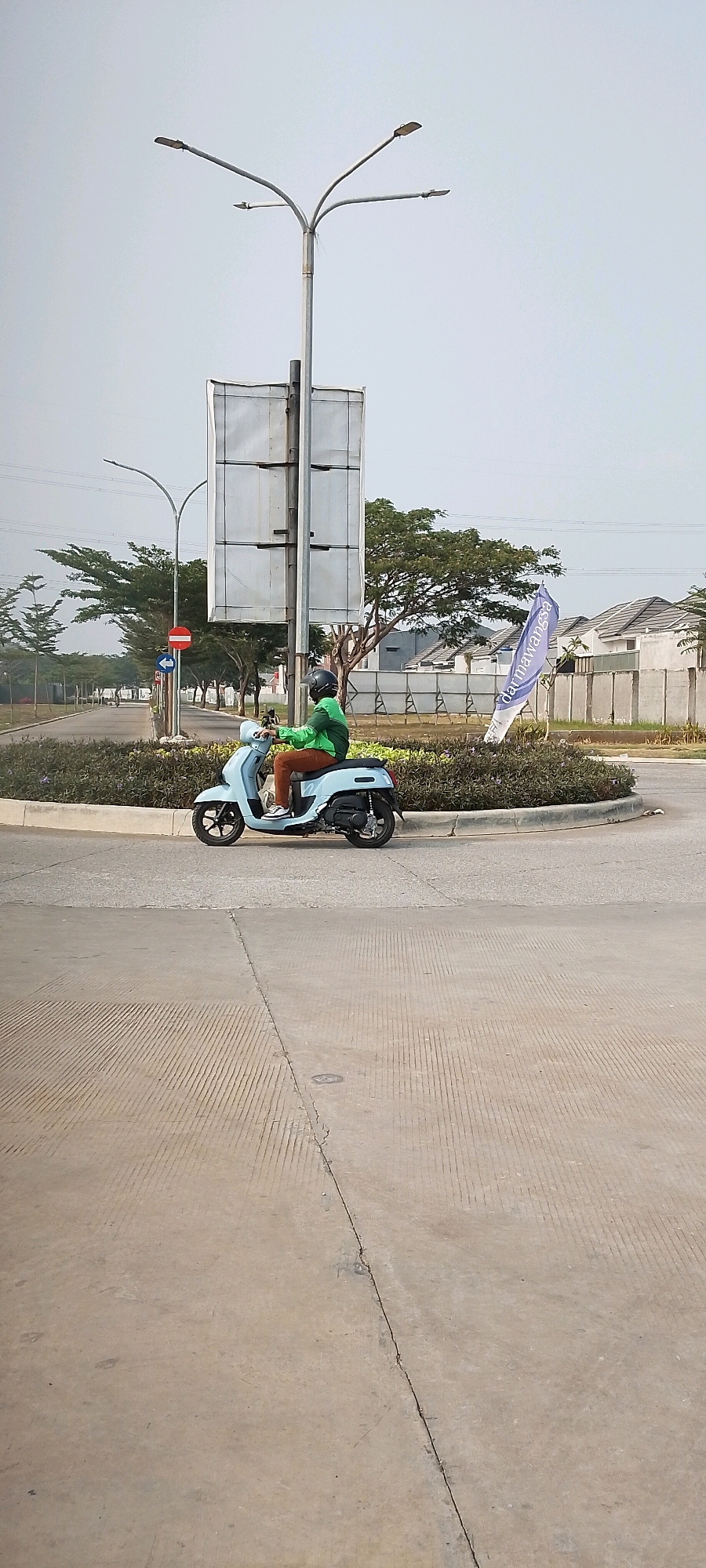 A person wearing a green jacket and brown pants rides a light blue scooter. The scooter has a black seat and black wheels. The rider wears a black helmet and white shoes. The scooter is positioned on a paved road with white markings, on the right side of the image. Behind the scooter, there is a circular garden with purple flowers and green bushes. A tall street lamp with four lights stands behind the garden. A large, rectangular white sign is attached to the pole of the street lamp. In the background, there are trees, houses with grey roofs, and a tall blue and white flag that reads "darmawangsa". The sky is overcast. The foreground of the image shows a concrete surface with textured lines running parallel to each other. The scene appears to be in an urban or suburban area, possibly a residential development given the houses and landscaping.