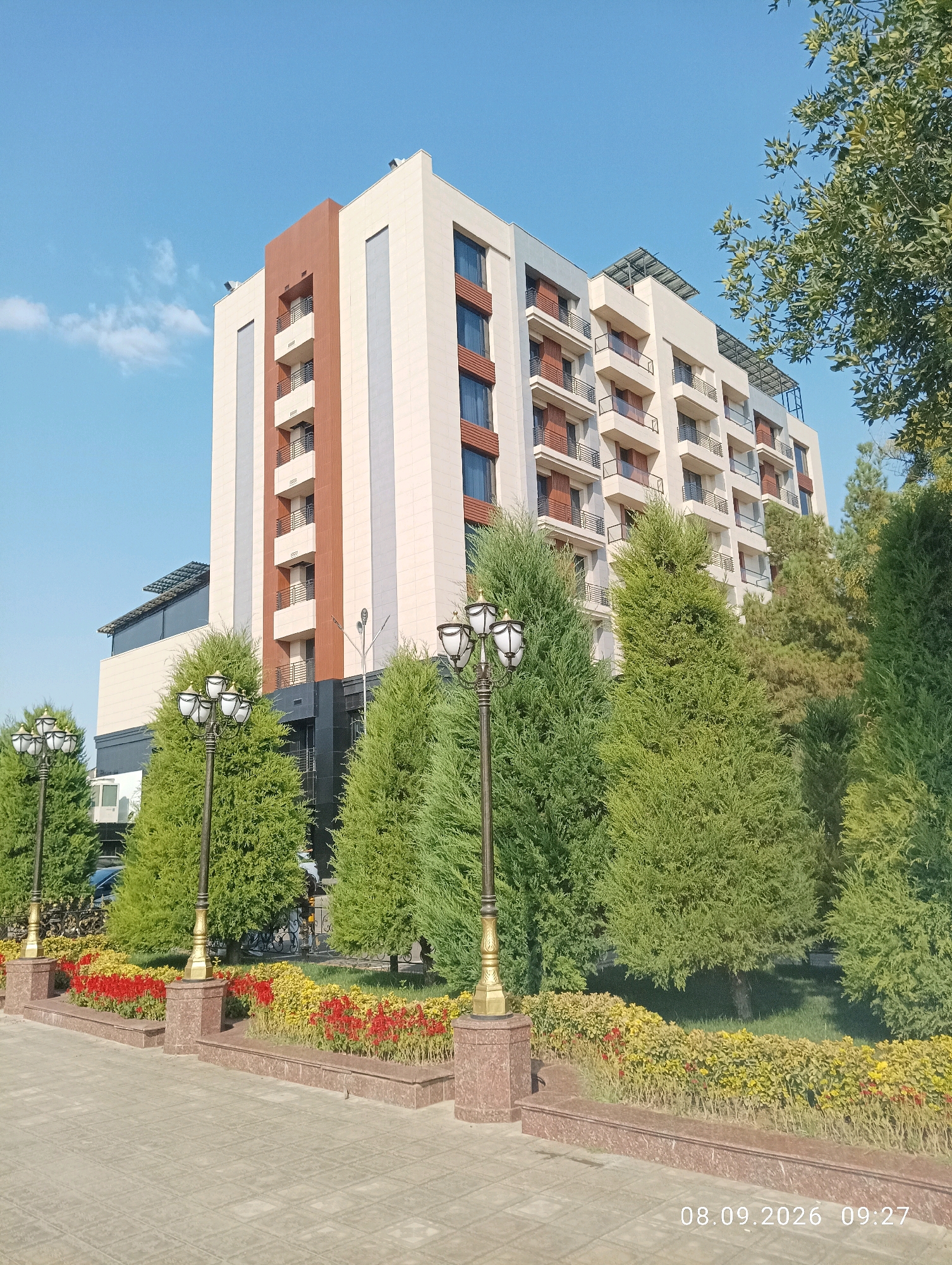 The image shows a modern, multi-story building with a light-colored facade, accented with brown and grey vertical stripes. The building has multiple balconies with dark railings. In front of the building is a landscaped area with manicured evergreen trees and flowerbeds containing red and yellow flowers. Several ornate lampposts with multiple lights are present along a paved walkway in the foreground. The sky is clear blue with a few wispy clouds. The date stamp at the bottom right reads "08.09.2026 09:27". This scene is consistent with the area around the Mirzo Ulugbek National University in Tashkent, Uzbekistan, specifically near the university's administrative buildings or dormitories. No people are clearly visible in the image.