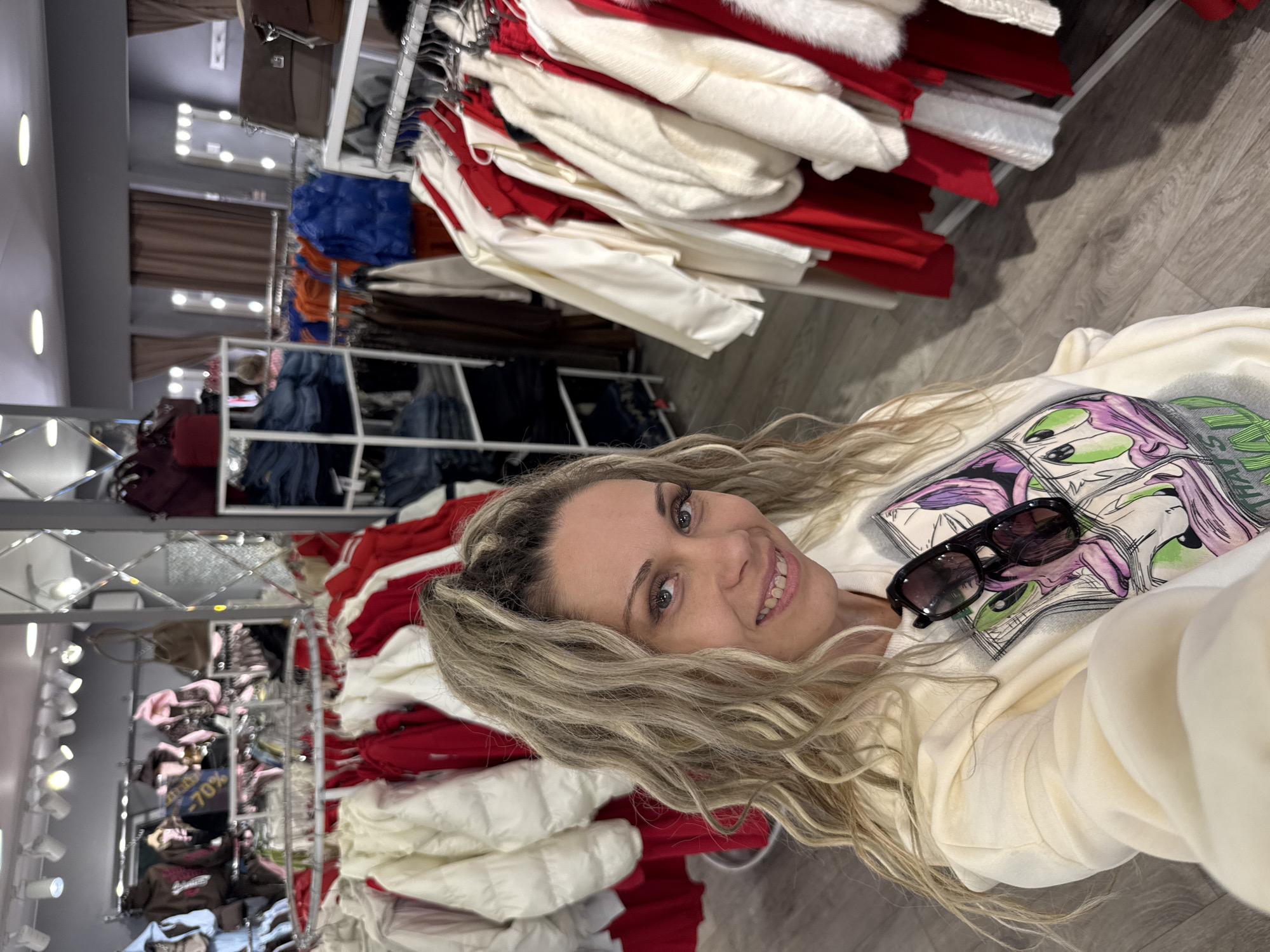 A woman with blonde, curly hair tied up in a ponytail is taking a selfie in what appears to be a clothing store. She is smiling and wearing a light-colored sweatshirt with a cartoon graphic on the front. A pair of sunglasses is perched on top of the sweatshirt. The background shows racks of clothing, including white and red jackets, as well as shelves with folded jeans. Mirrored panels and light fixtures are visible on the ceiling and walls. The flooring is light wood. There is no information that allows for the location to be determined as Rusanovo, Russia.