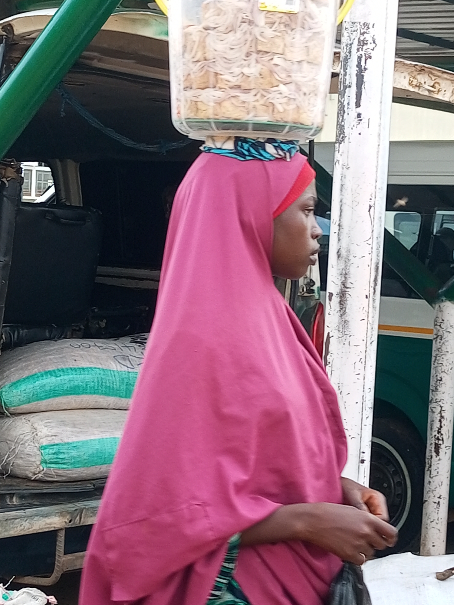 A person wearing a pink headscarf and pink garment is walking outdoors in a city, possibly Gombe, Nigeria, carrying a clear plastic container filled with food items on their head. The person is walking next to a vehicle with a green stripe, and in the background, there are more vehicles and structures. In the foreground, there are two large sacks, one with a green stripe. The person's hands are visible, and they are holding a dark bag.