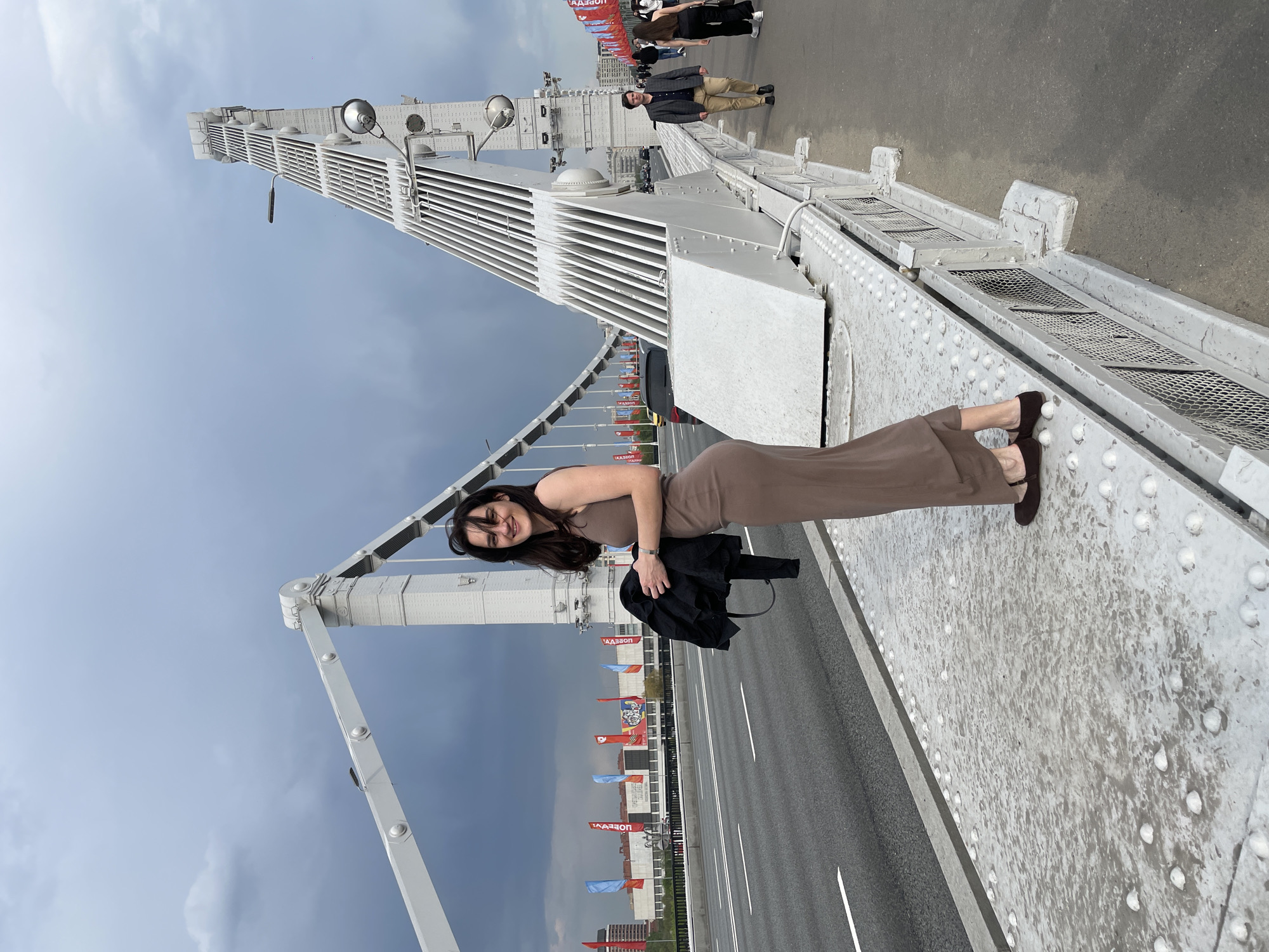 A woman is standing on a pedestrian walkway of the Krymsky Bridge, Moscow, Russia. She is wearing a light brown dress and brown shoes. She is holding a black jacket in her left hand. The bridge's architectural details are visible, including the white metal framework and rivet-covered surfaces. In the background, there are flags and other people on the bridge. There is a highway beneath the bridge. The sky is overcast.