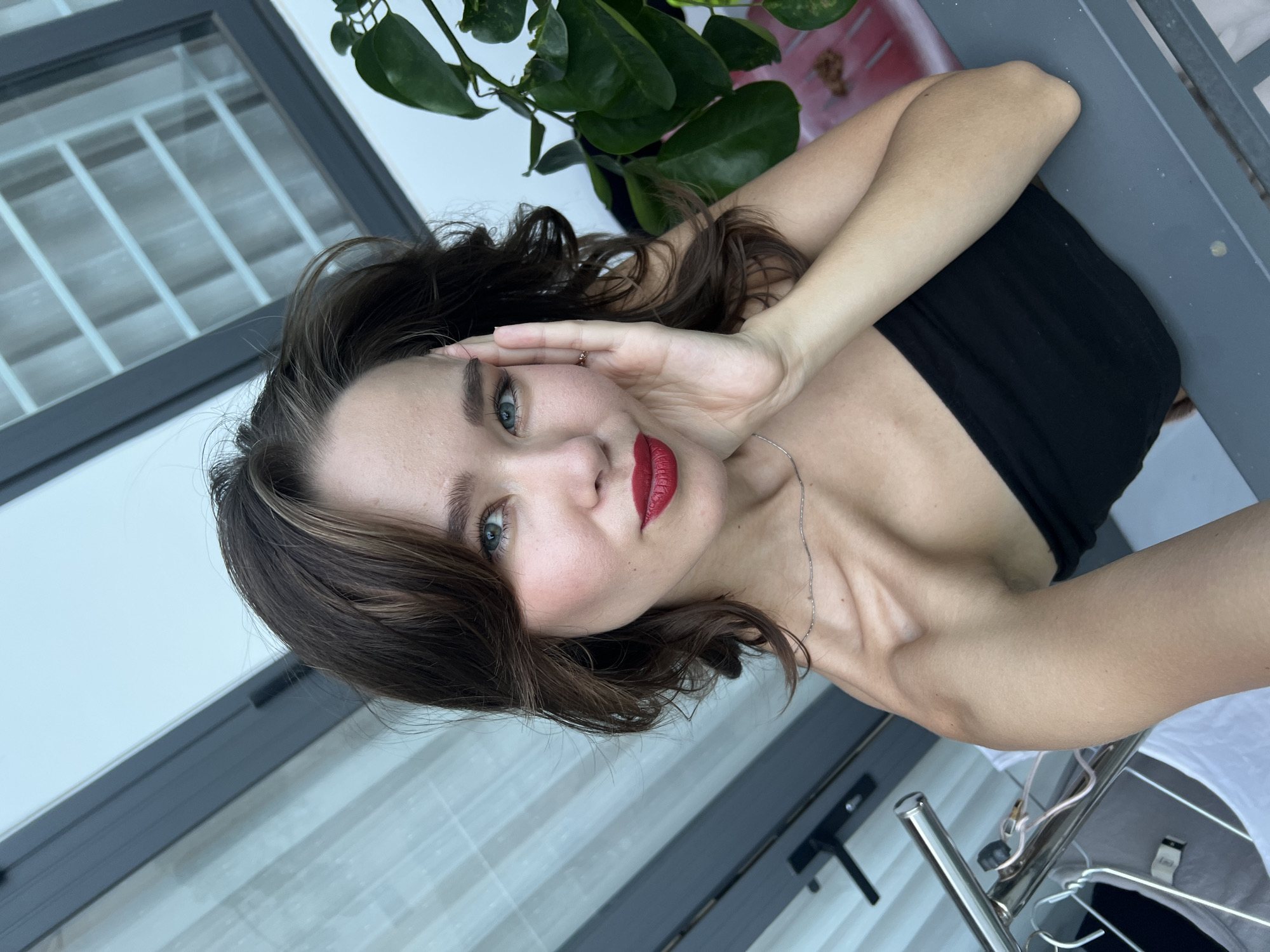 The image shows a woman with long brown hair and bright red lipstick posing for a photo. She is wearing a black strapless top and a silver necklace. Her right hand is raised near her face, and she is looking directly at the camera. The background includes a window with white panes and a potted green plant. A gray, possibly metal, structure is also visible in the foreground.
Based on the location information (Nam Nha Trang Ward, Vietnam), the scene is likely an outdoor or semi-outdoor space such as a balcony or terrace in a residential or commercial building.