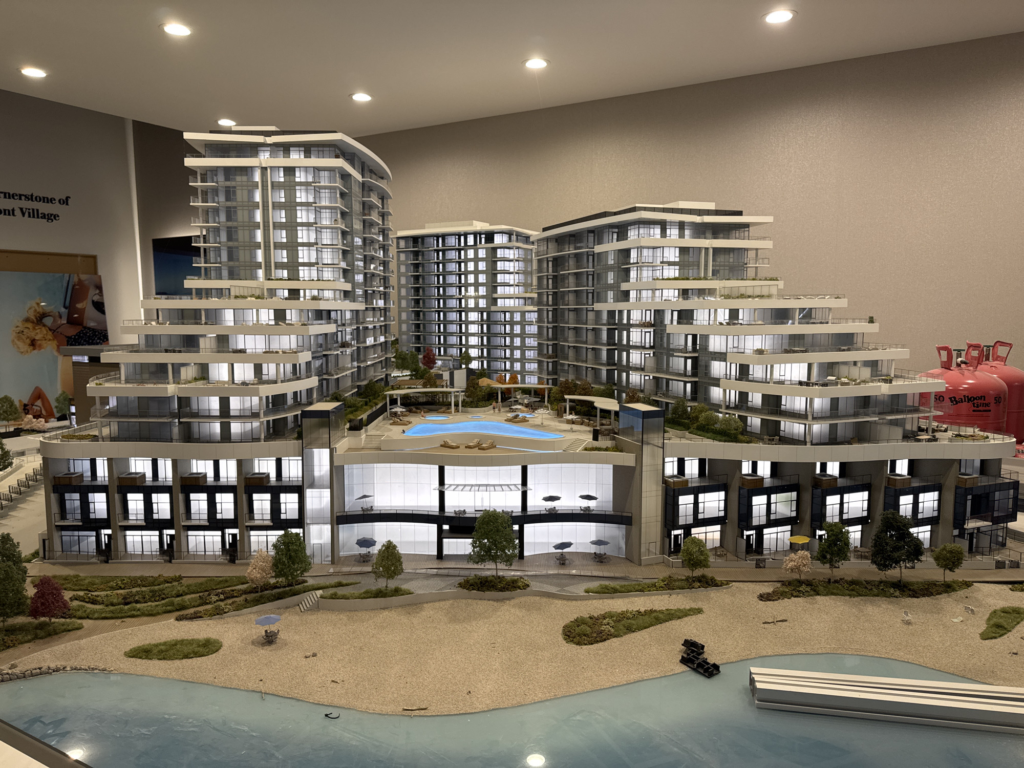 This image is a detailed architectural model of a waterfront residential complex. The model depicts multiple buildings with a tiered, stepped design, featuring balconies, large windows, and modern architectural elements. The complex includes amenities such as a swimming pool, lounge chairs, umbrellas, and landscaped green spaces, all situated on a sandy beach area adjacent to a body of water. The model is lit from within, simulating interior lighting in the residential units. In the background, part of a poster with text is visible, and to the far right, two large red propane tanks are present. The scene is viewed from a slightly elevated perspective, showcasing the scale and layout of the development. The location is identified as Kelowna, Canada, suggesting this is a model for a real estate development project in that city.