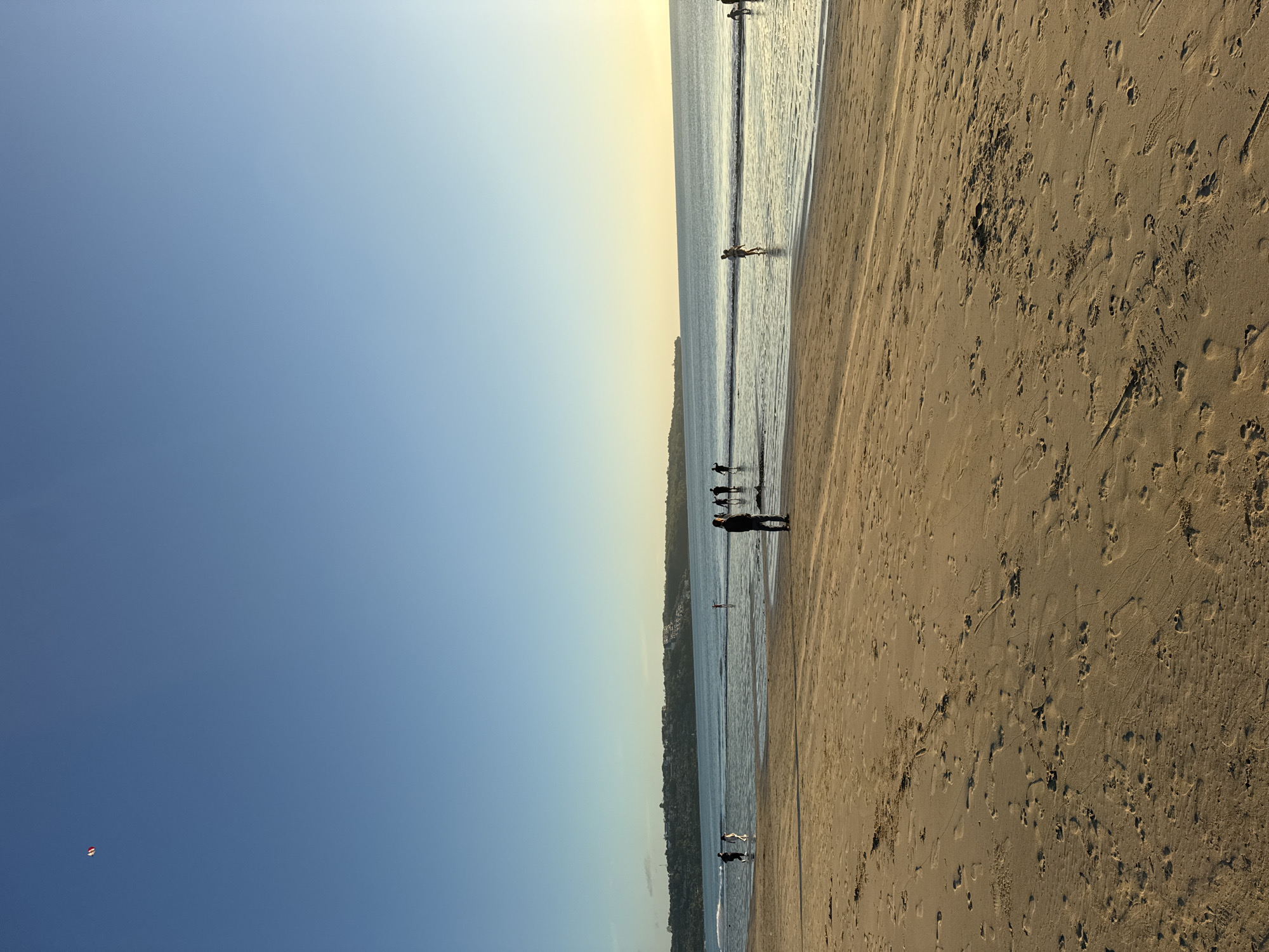 The image depicts a wide, sandy beach in Kuta Selatan, Indonesia, during what appears to be late afternoon or early evening. The sky is a clear, gradient blue, transitioning to a pale yellow near the horizon where the sun is setting. The ocean is calm, with gentle waves lapping the shore. Several people are visible on the beach and in the shallow water. In the foreground, footprints and tire tracks are imprinted in the wet sand. Further back, a cluster of individuals stands near the water's edge, some wading, while others are closer to the shore. A distant coastline with lush vegetation and buildings forms the backdrop. In the upper left quadrant of the sky, a small, colorful kite is visible. The overall scene conveys a sense of peaceful recreation.
