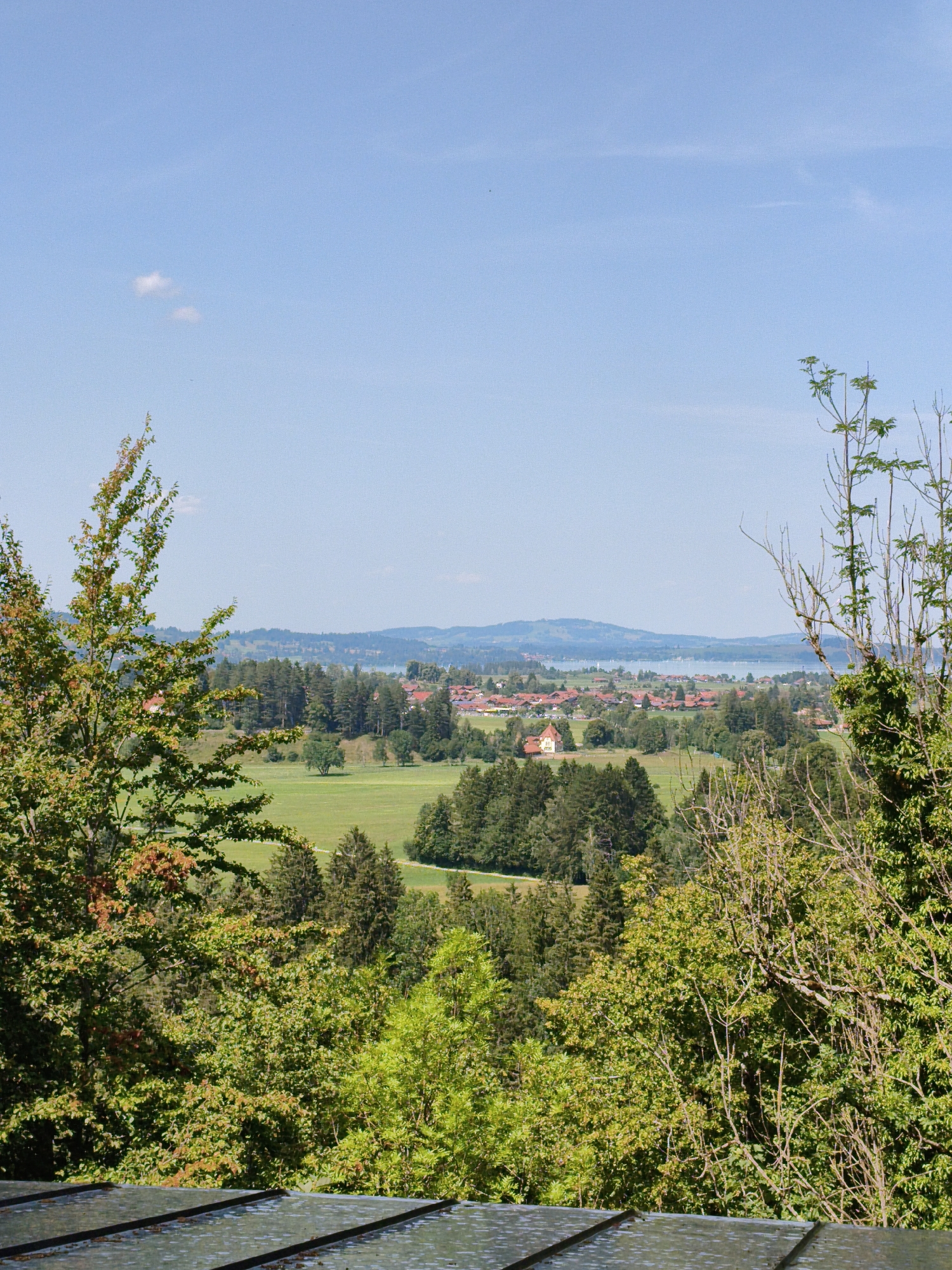 The image depicts a panoramic view of a town nestled amidst rolling green hills and a body of water, with a clear blue sky overhead. In the foreground, a textured, dark surface, possibly a roof or deck, is visible. The middle ground is dominated by lush green trees and fields, with scattered houses featuring red-tiled roofs. In the distance, a town stretches along the shoreline of a lake, with hills forming a backdrop. The overall scene suggests a tranquil, rural setting, characteristic of the Bavarian Alps region. While the specific location within Schwangau, Germany cannot be pinpointed from the image alone, it is representative of the picturesque landscapes found in the area, which is renowned for its natural beauty and proximity to famous castles. There are no people or animals visible in the image.