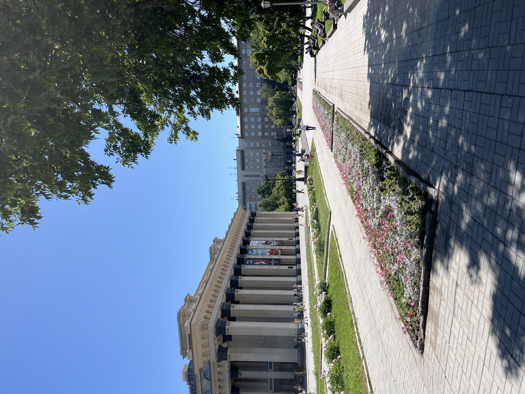 An outdoor, eye-level, full shot captures a bright, sunny day in a city park. A large, classical building with numerous tall, stone columns dominates the left side of the frame. Banners are hung between the columns. People are walking along the sidewalk in front of the building. In the foreground, a wide, paved walkway stretches into the distance, flanked by a long, manicured flowerbed filled with pink and white flowers. To the right of the walkway, a section of grass with benches and people seated on them is visible. Beyond the flowerbed, another paved path leads towards a residential building. Lush green trees, including one prominent tree in the upper right corner casting shadows on the pavement, provide shade. The sky is a clear, vibrant blue. The scene appears to be a public space designed for recreation and cultural engagement.