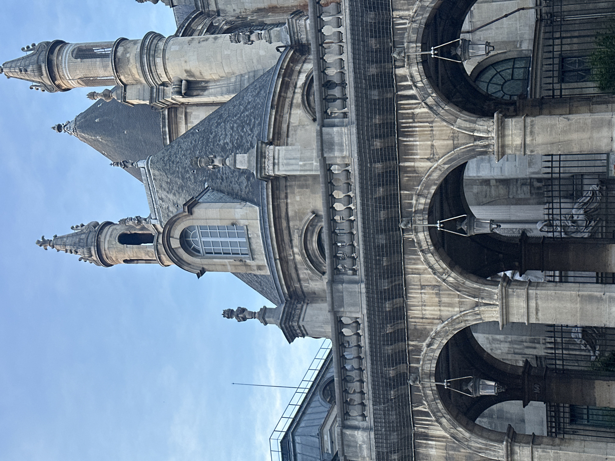The image displays a detailed view of a grand, ornate stone building, characteristic of historical architecture found in Paris, France, set against a clear blue sky. The architectural style appears to be classical or neoclassical with Baroque elements, featuring grey stone and intricate sculptural details.
In the lower and mid-sections, the building presents a series of large, rounded arched openings, possibly leading to an interior or forming a loggia. Decorative street lamps hang within some of these arches. One prominent stone pillar within an arch clearly shows the etched number "160". Behind the arches, parts of dark wrought-iron gates or grilles are visible.
Higher up, the structure features steep, dark slate roofs, interspersed with dormer windows that have arched tops and multi-pane glazing. The building's facade is adorned with ornate cornices, balustrades, and numerous decorative elements including finials and sculptural forms along the roofline and various ledges.
Two slender, highly decorative spires or turrets rise prominently towards the sky. Each spire is topped with an elaborate finial and features openings—one with a round arched window below it, and another with tall, narrow, arched openings.
The natural light suggests it is daytime, with bright, direct sunlight illuminating the stone and creating subtle shadows, indicating fair weather conditions. No people or specific activities are visible, focusing solely on the architectural grandeur.