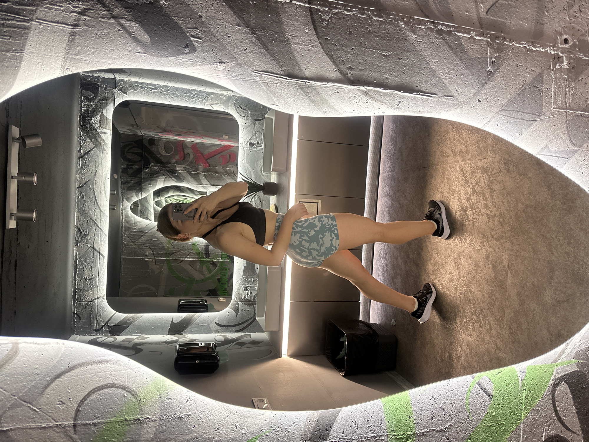 The image captures a young woman taking a mirror selfie in a modern, urban-style restroom. She is wearing a black sports bra and light blue camouflage-patterned shorts, along with black athletic shoes. The restroom features a large, irregularly shaped mirror, which reflects the woman and the surrounding environment. The walls of the restroom are decorated with graffiti-style art in shades of gray, black, and green, giving it a gritty, industrial aesthetic. There are modern bathroom fixtures, including white sinks with chrome faucets, and overhead lighting. The floor is a neutral-toned tile. The image does not provide any information that would allow for identification of the city as Ternopil, Ukraine.