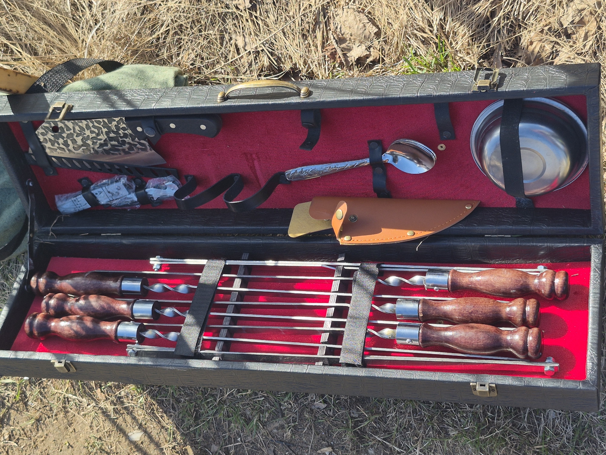 The image shows an open black suitcase with red lining, containing various cooking and grilling utensils. The suitcase is resting on a grassy area with dry leaves and some green vegetation.
The top compartment of the suitcase contains a meat cleaver in a patterned cover, a first aid kit, a long-handled spoon, a knife with a leather sheath, and a stainless steel bowl secured with a black strap.
The bottom compartment holds metal skewers with wooden handles, organized by black straps. The skewers appear to be for grilling kebabs.
There are no people visible in the image. The location is likely outdoors, suggested by the ground cover, but the city of Chaikovsky, Russia is not ascertainable from visual elements of the photograph.