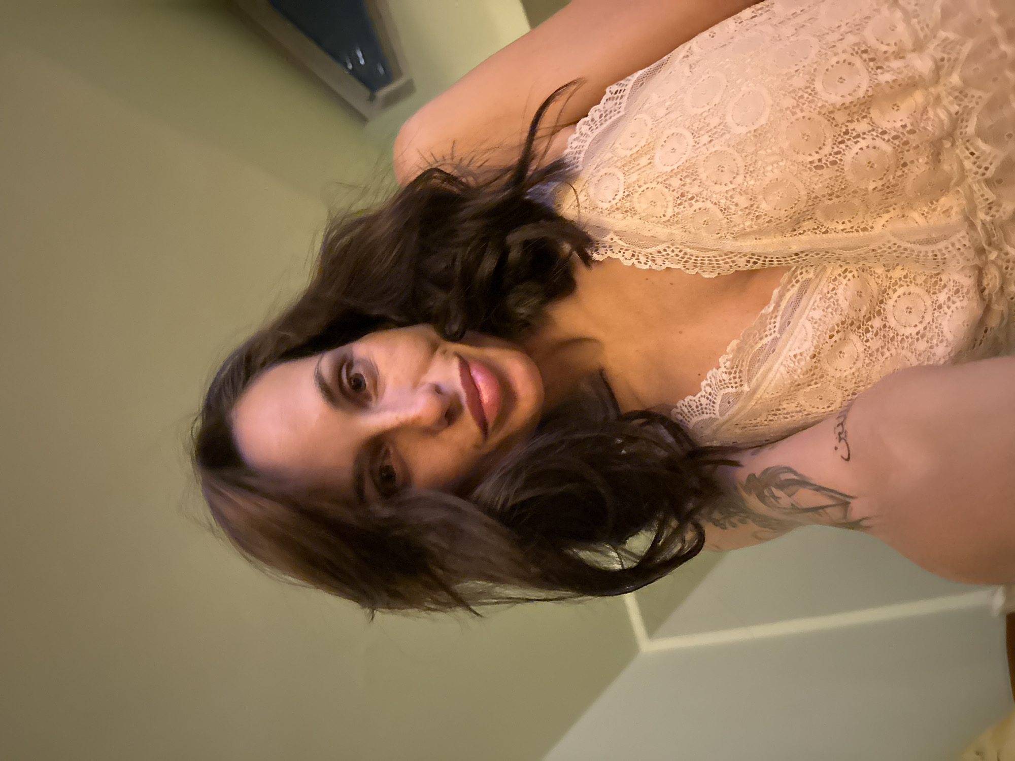 A medium shot depicts a woman with long, dark hair, fair skin, and brown eyes. She is wearing a lacy white top. A tattoo is visible on her left arm. The background consists of pale green walls and white trim. The location is unknown.