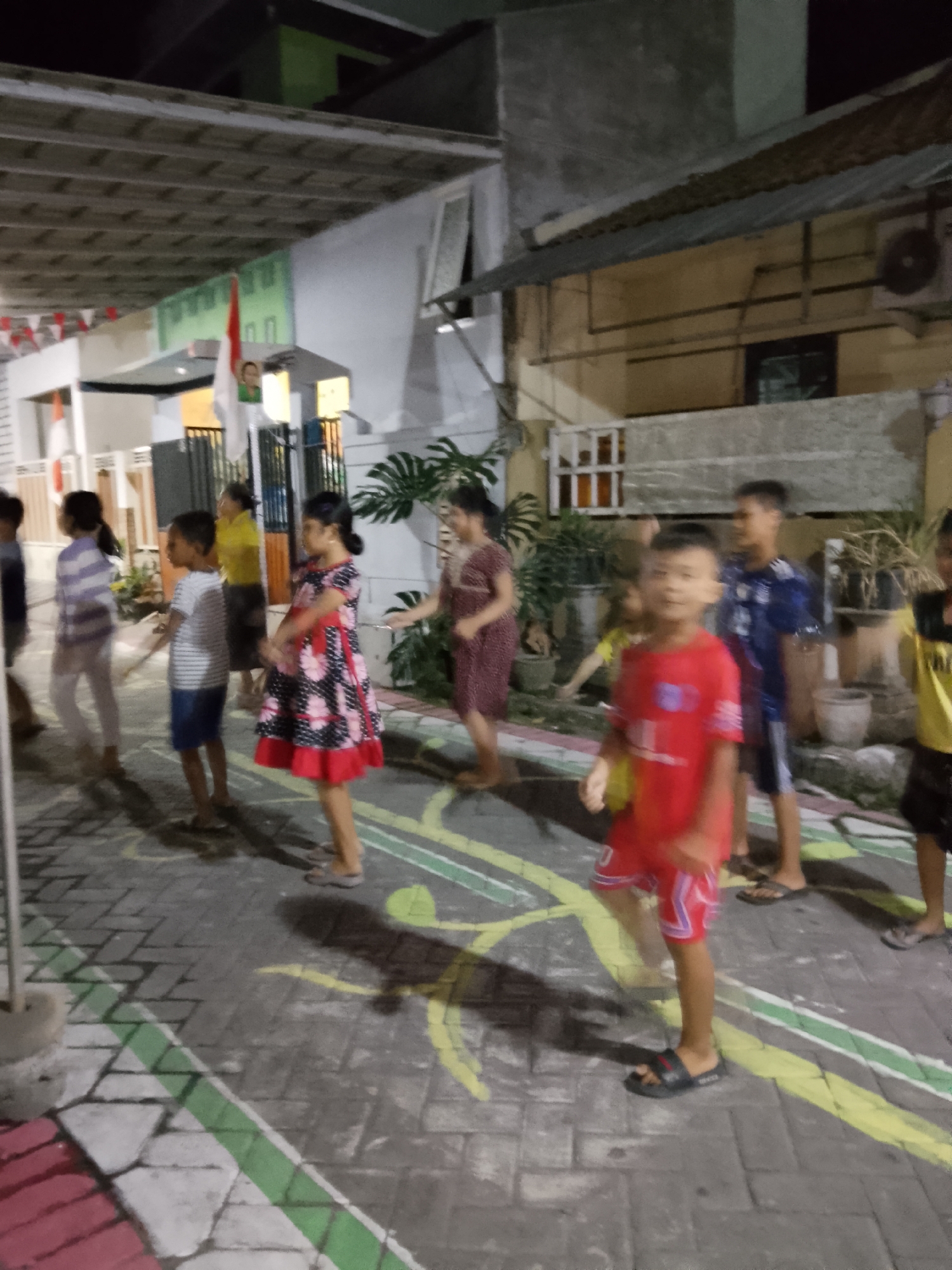 In Banjarsari, Indonesia, a group of children are gathered on a paved outdoor area that appears to be a street or alleyway. The ground is made of interlocking grey and white brick pavers, with painted yellow and green lines forming what could be game markings or directional arrows. Several children are visible, most in motion. In the foreground, a boy in a red shirt and red shorts with white trim is running or dancing, wearing black slide sandals. To his left and slightly behind him, a girl in a black and red floral dress is also in motion, her left arm extended. Further back, other children are seen in various states of activity. One child wears a striped shirt and blue shorts, another a purple and white striped shirt with light-colored pants, and a girl in a yellow shirt is in the background. The scene is lit by artificial light, casting shadows on the ground. In the background, buildings are visible, with a slanted corrugated metal roof and part of a white wall with a window. A flag with red and white stripes and a portrait on a pole is also present. The overall impression is one of an informal gathering or activity taking place in a residential or communal outdoor space.