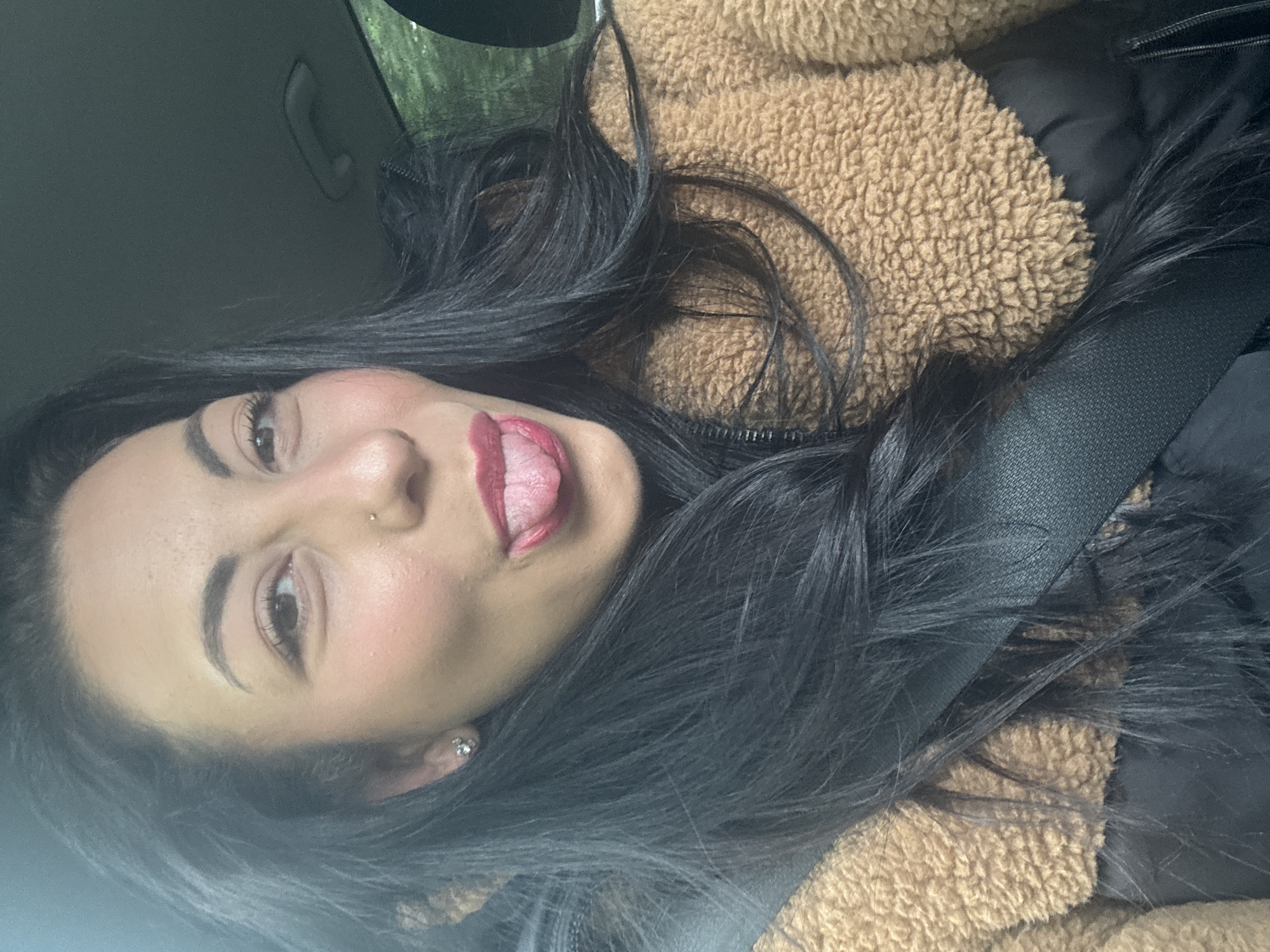 A young woman is in the driver's seat of a car, possibly in Warrington, UK, though the exact location is not evident from the image. She is wearing a seatbelt and a tan, textured jacket over a black garment. Her dark hair is styled loosely around her face. She has pierced ears, a nose ring, and is sticking her tongue out. The interior of the car is visible, with the car door and possibly a part of the exterior visible through the window.