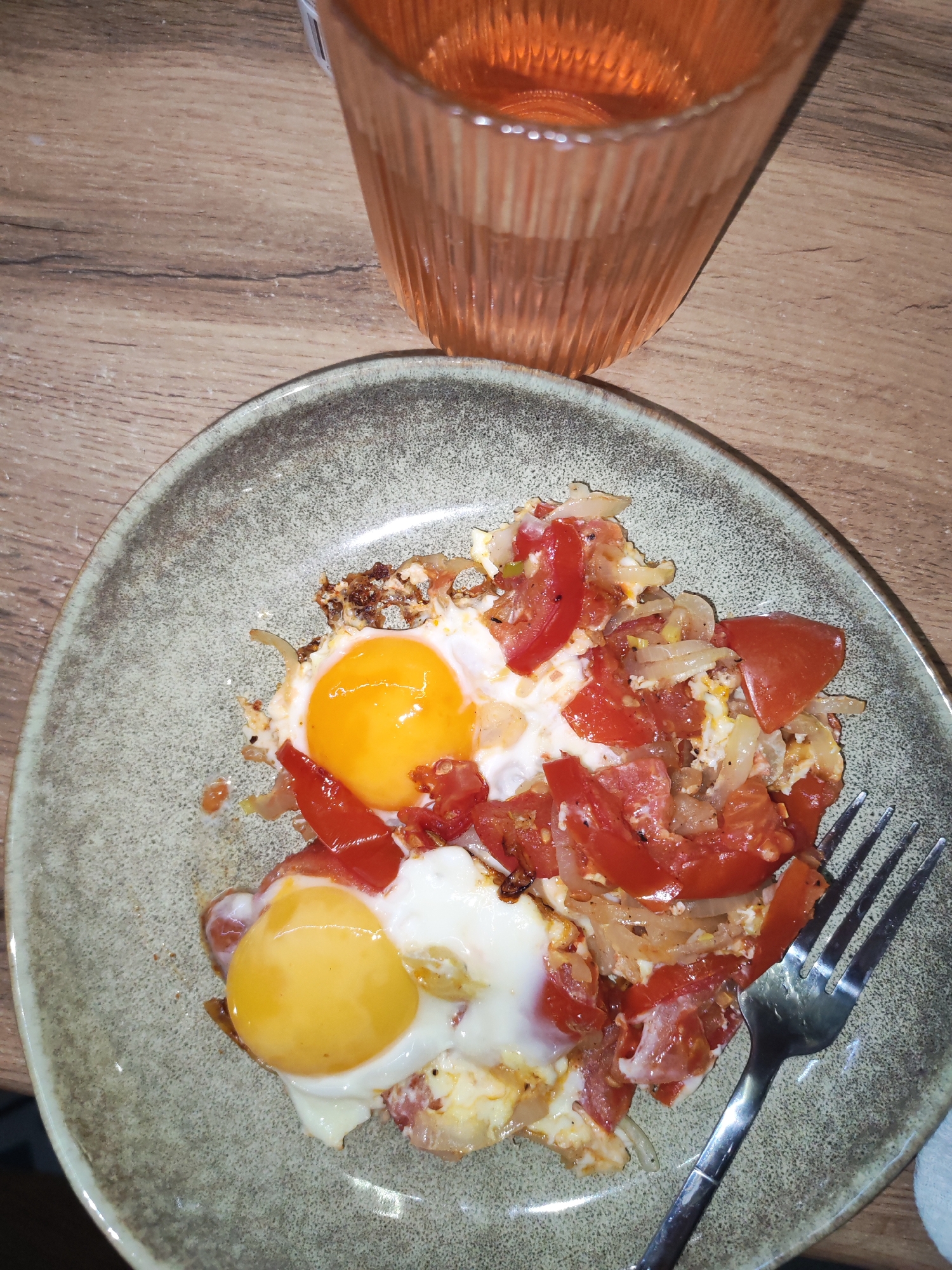 The image shows a plate of food on a wooden table, with a glass of liquid nearby. The food appears to be fried eggs with tomatoes and onions. A fork rests on the plate. The glass contains a light orange-colored liquid, possibly juice or wine. The scene takes place on a wooden surface indoors. There is no indication of the location being Izhevsk, Russia.