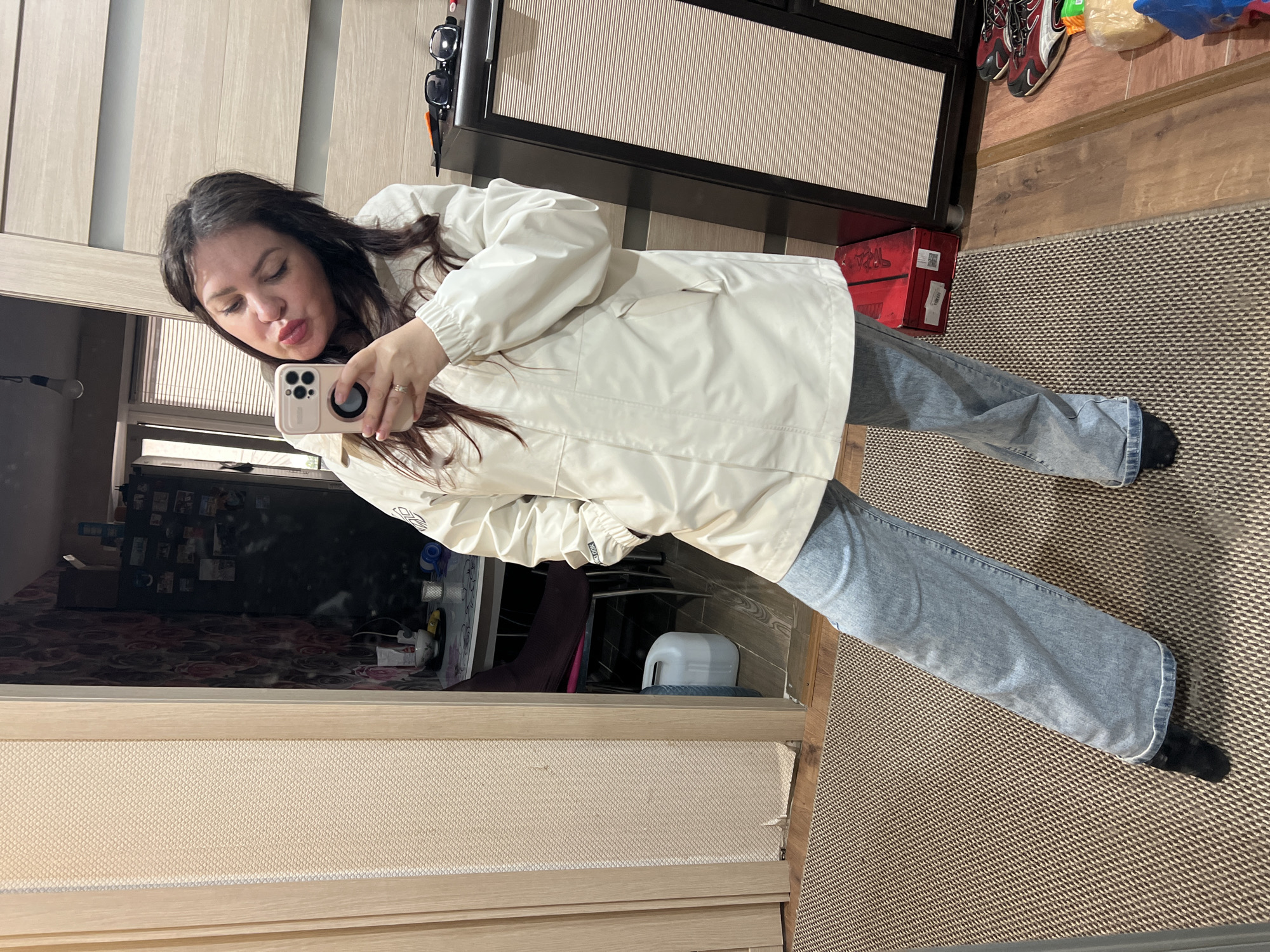 A young woman is captured in a mirror selfie taken indoors in Noviy, Russia. The image is oriented sideways, showing her standing with her body angled towards the camera, holding a light pink or beige smartphone in her right hand, which has a triple camera setup. She has long, dark brown hair and is making a "pouty" or "kissing" expression towards her reflection.
She is wearing a light cream or off-white long-sleeved jacket or coat and light-wash blue jeans. Her feet, clad in black socks, are visible at the bottom of the jeans. On her right ring finger, a ring is visible.
The setting appears to be a casual home environment. The woman is standing on a patterned rug with a brown and off-white geometric design, adjacent to wooden or laminate flooring. In the reflection behind her, a dark cabinet or wardrobe with light-colored, possibly slatted, panels is seen. On top of this furniture, a pair of dark sunglasses rests. Below it, a red box with partially visible text, possibly "TRPA" or "TRAP", is on the wooden floor, next to what appears to be a pair of dark red and black athletic shoes. A plastic bag is also discernible in the background. Other reflections show parts of a room with different flooring patterns and various household items, including a white plastic container.