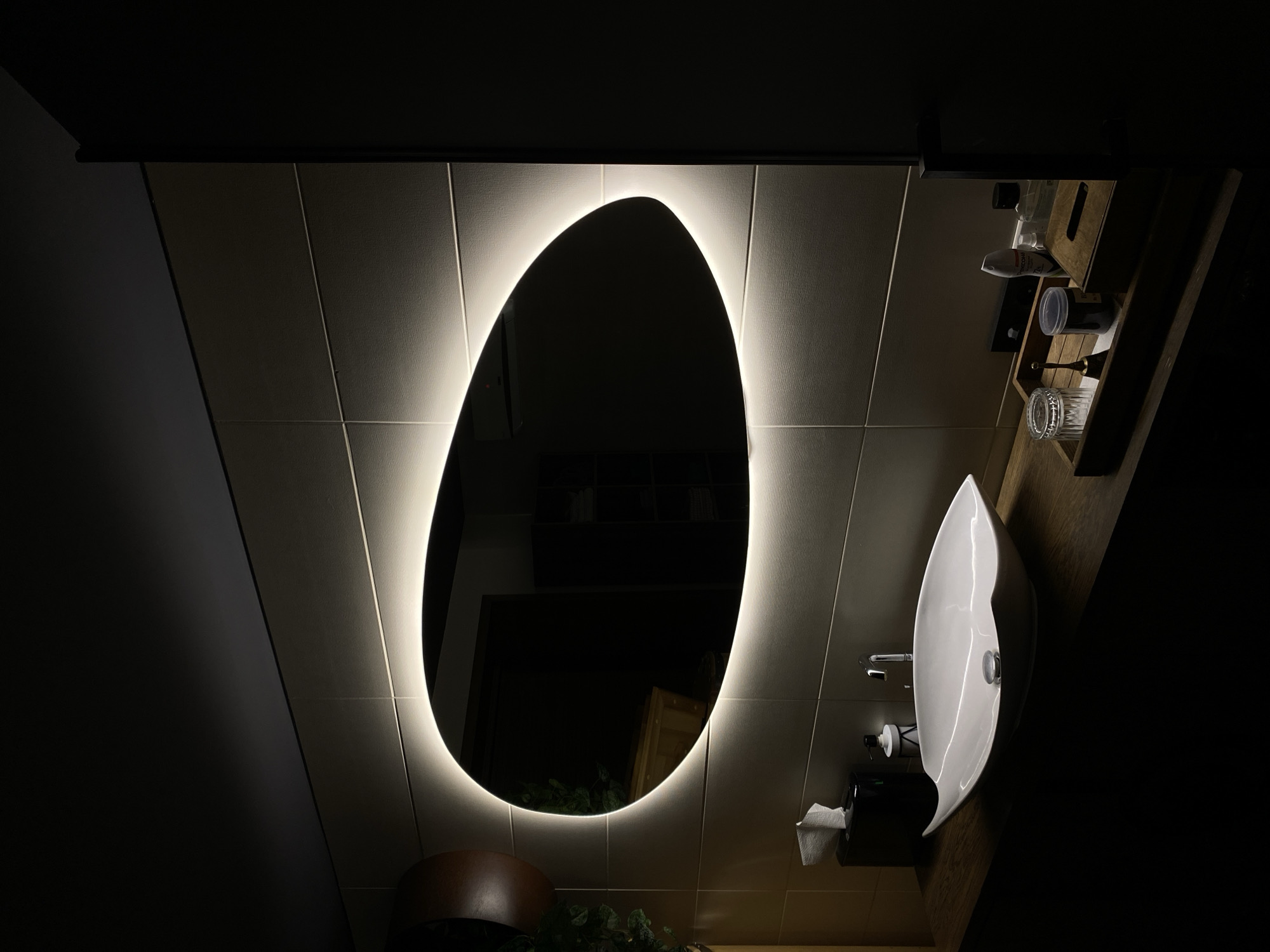 The image depicts a dimly lit bathroom. The main focus is an irregularly shaped mirror with a bright, glowing outline, mounted on a tiled wall. The tiles are light-colored with visible grout lines. A white, teardrop-shaped sink with a chrome faucet is situated to the right of the mirror. A dark-colored counter supports the sink. A tissue dispenser and other bathroom accessories are positioned near the sink. A shelf with toiletries is mounted on the wall above the sink. The lighting is such that the right side of the image is almost entirely dark, creating a high contrast. The reflection in the mirror shows a room with shelves. It is unlikely this image is in Votkinsk, Russia.