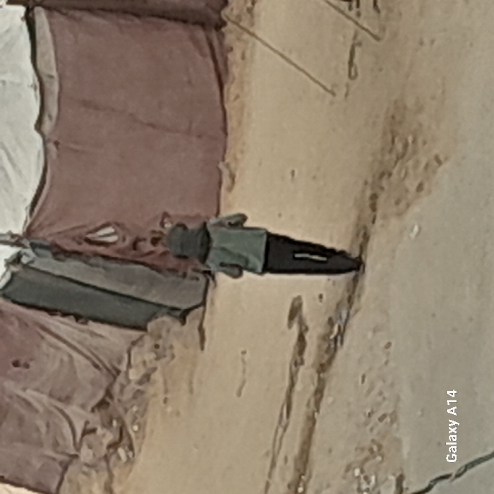 A person in a green shirt and dark pants lies on the ground next to some objects covered with a maroon cloth, beside a sandy surface. The scene is captured with an "Galaxy A14" phone watermark appearing in the lower right corner. The overall image has a stylized or edited appearance.