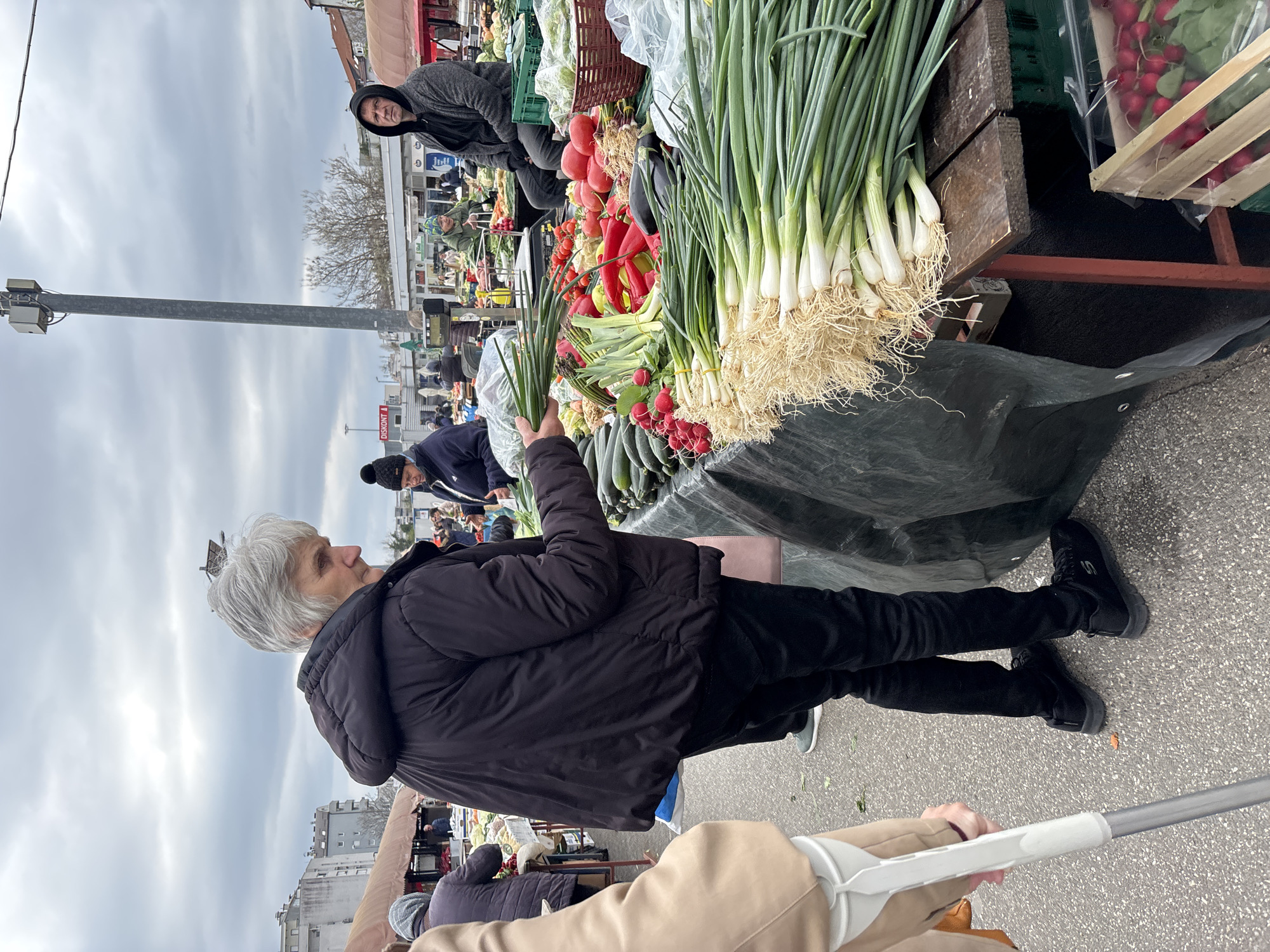 An eye-level, outdoor shot captures a bustling marketplace in the city of Zagreb, Croatia. A woman with short gray hair, wearing a black jacket and black pants, stands at a produce stall, holding a bunch of green onions. The stall is laden with fresh vegetables, including green onions, tomatoes, peppers, zucchini, and radishes. A man in a gray hoodie sits behind the stall. Other shoppers and stalls are visible in the background. The weather appears overcast. A walking stick is partially visible in the foreground. A tall light post can be seen in the background of the left side of the shot. There are buildings in the far background of the left side of the shot.