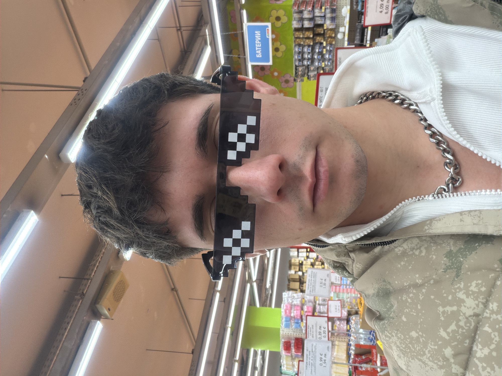 A young man wearing pixelated sunglasses and a silver chain is standing inside what appears to be a supermarket or a general store. The location is suggested to be Sofia, Bulgaria due to the Cyrillic script on signs, specifically the word "БАТЕРИИ" (batteries). The man is wearing a camouflage jacket over a white hoodie. The background shows shelves filled with various products and price tags in Euros and Bulgarian Levs. Fluorescent lights are visible on the ceiling.