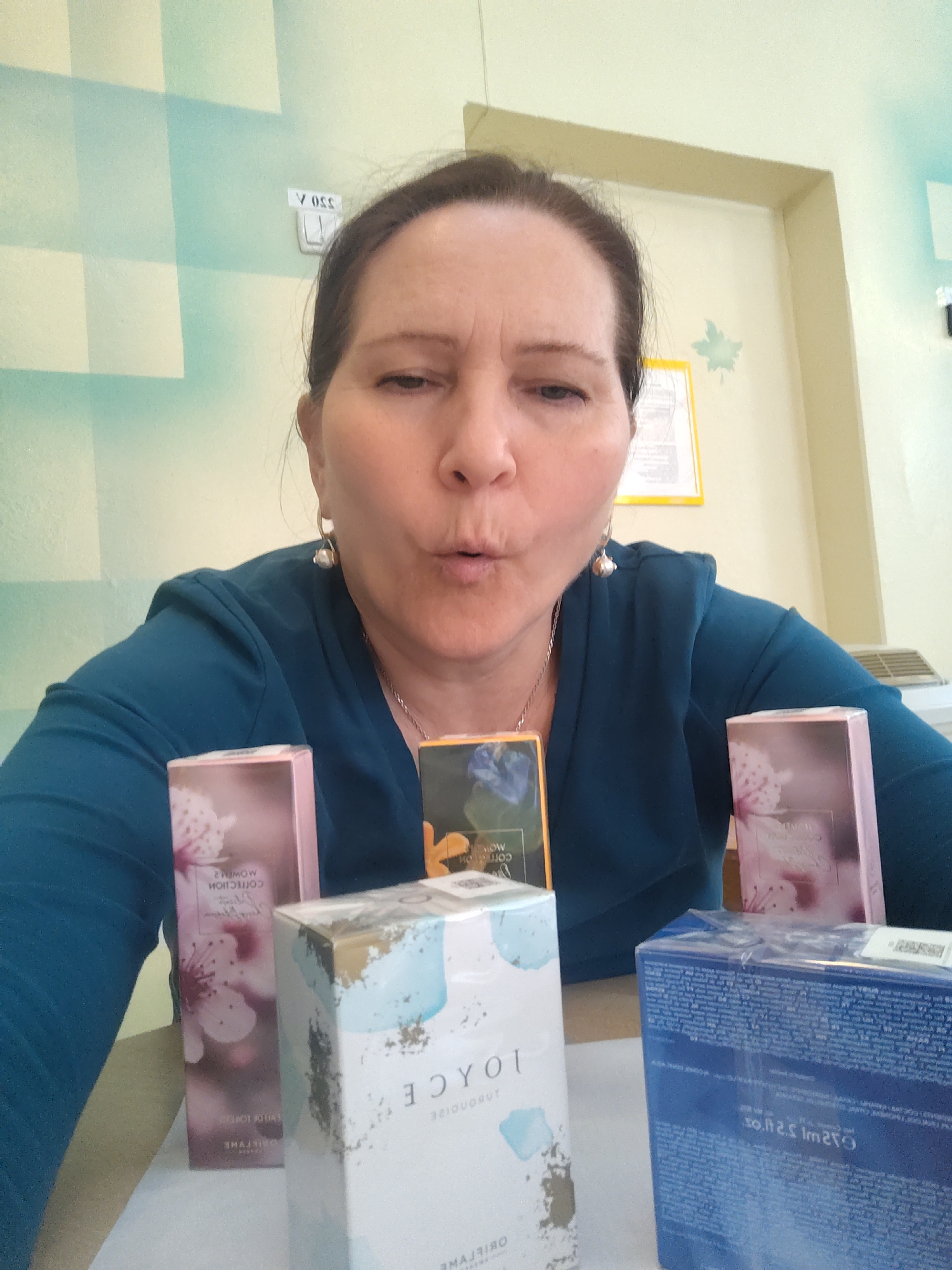 A woman with brown hair pulled back, wearing a teal top and silver earrings and necklace, is holding several perfume boxes. The boxes are of varying designs and include the brand "Oriflame". The woman is making a "kissing" face towards the camera. The background includes a wall with a light-blue and cream-colored checkered pattern, a doorway, a yellow-framed notice, and what appears to be a heating/cooling unit. The location is likely Sarapul, Russia, given the Oriflame brand's presence in that region.
