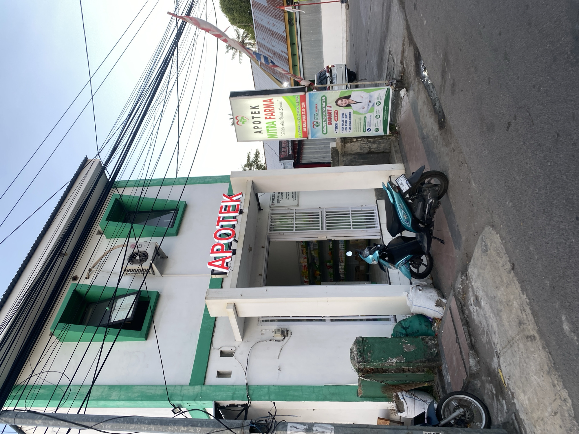 This is a ground-level, eye-level, medium shot of the exterior of a small, white and green pharmacy in Bantaeng, Indonesia. The pharmacy building has a modern facade with green trim around the windows and roofline. Above the entrance, the word "APOTEK" is displayed in large red letters. A signpost to the right of the entrance displays "APOTEK MITRA FARMA" and information about health consultations. A turquoise scooter is parked to the left of the entrance, and a dark blue scooter is parked further left. A white SUV is parked on the street behind the signpost. The sky is clear and blue, with several power lines crisscrossing the frame. The overall impression is of a typical urban street scene in Indonesia.