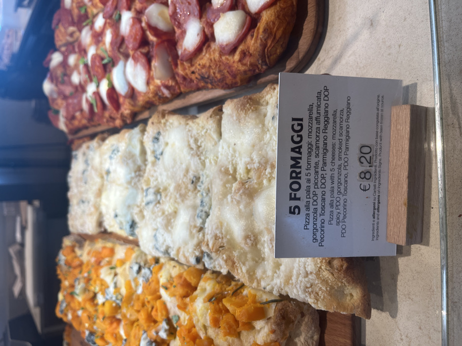 This image captures a vibrant display of various rectangular pizzas or focaccia bread, likely presented for sale in a food counter or a bakery/deli in Dorno, Italy. The pizzas are arranged on wooden serving boards, creating an appetizing visual.
Three distinct types of pizza are visible. At the top, a pizza is generously topped with slices of pepperoni or salami and white cheese chunks, resting on a golden-brown, thick crust. Below it, a "5 FORMAGGI" (Five Cheese) pizza features a creamy white cheese topping with visible blue veining, indicative of gorgonzola, alongside other white cheeses. At the bottom, another pizza showcases an orange-colored topping, possibly roasted butternut squash or sweet potato, garnished with green herbs.
A white information card is prominently displayed in front of the five-cheese pizza. The card, written in both Italian and English, specifies the ingredients for the "5 FORMAGGI" pizza: "mozzarella, spicy PDO gorgonzola, smoked scamorza, PDO Pecorino Toscano, PDO Parmigiano Reggiano." The price for this item is clearly marked as €8.20. The card also notes that ingredients and allergens are available on separate signs, and the product is made from a frozen base.
The scene suggests an indoor commercial setting during business hours, with the prepared food ready for purchase. No individuals are visible in the frame, and the time of day is not ascertainable. The close-up perspective emphasizes the textures and toppings of the different pizza varieties.