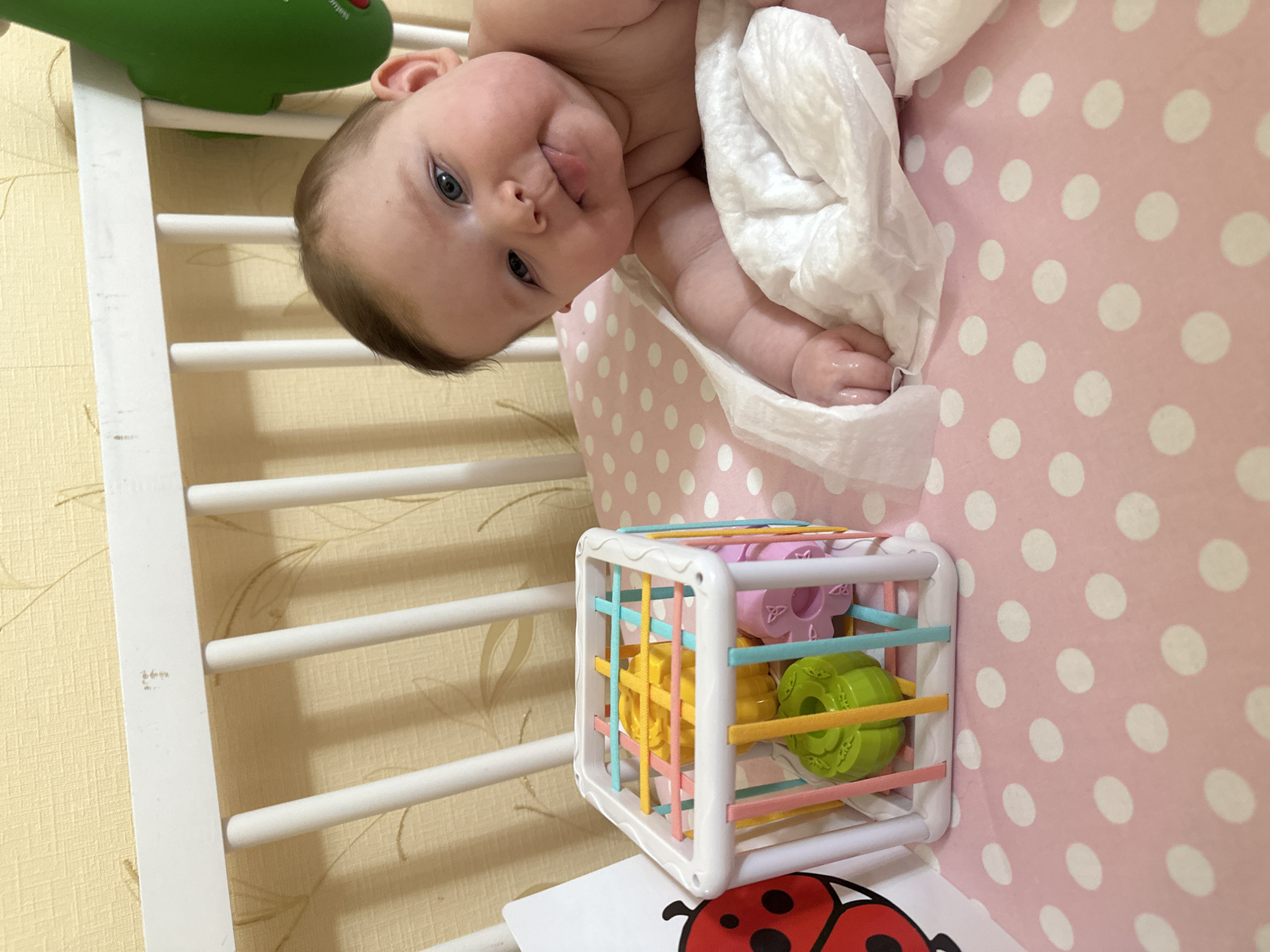 The image depicts a baby lying on their back in a crib. The baby is wrapped in a white cloth and has their tongue slightly sticking out. The crib has white rails and a pink polka-dot patterned liner. A white toy cube with colorful shapes inside is visible near the bottom of the image. A card with a ladybug illustration is partially visible under the toy cube. The crib is situated in a room with patterned wallpaper.
The image was taken in Chaikovsky, Russia.