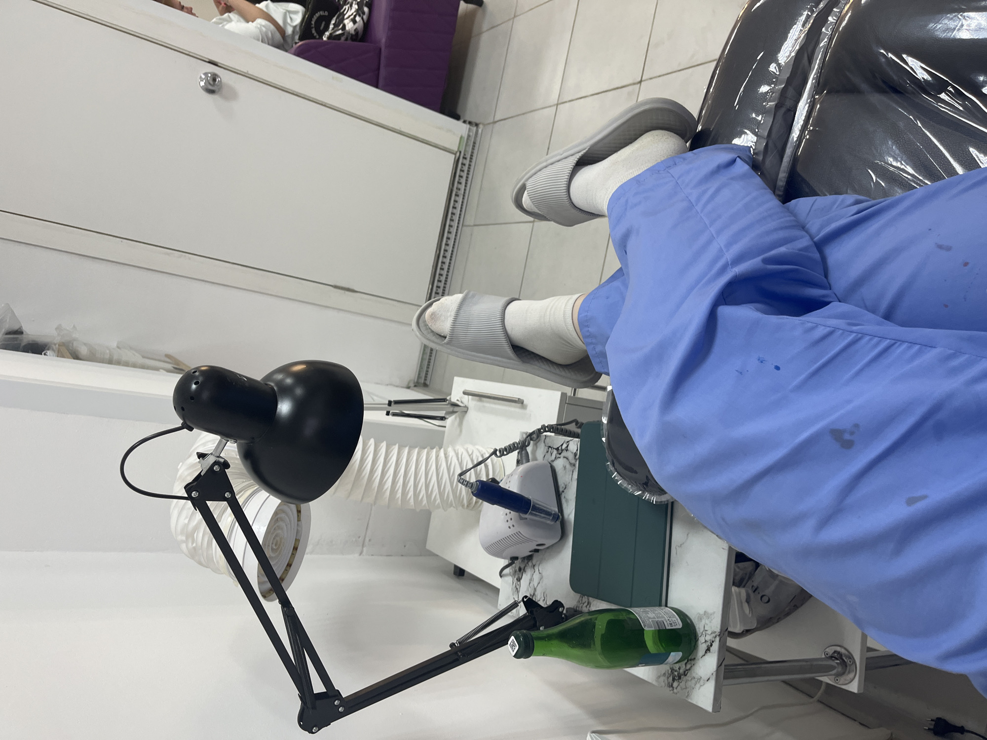 An indoor scene, rotated 90 degrees clockwise, depicts a professional setting, likely a nail salon, in Perm, Russia.
In the foreground, a person's legs are visible, dressed in light blue scrubs or trousers and grey, ribbed slippers over white socks. The blue attire has several dark splatters or stains. The legs rest near a black chair or stool covered in clear plastic.
Adjacent to the person's legs is a white table with a marbled top, which serves as a nail workstation. On the table, a black adjustable desk lamp is positioned. Also present are a white electric nail file machine with a blue drill bit or handle attached, and a dark green tablet case. A green glass bottle, possibly for nail polish remover or other liquid, lies on its side, bearing a small white barcode sticker near its cap. A white flexible tube, indicative of a dust extractor or ventilation system, extends from the workstation.
In the background, a client is partially visible, reclining on a piece of furniture, possibly a bed or comfortable chair. They are wearing a white t-shirt, with only their upper torso and head visible. Behind the client, a purple upholstered chair or sofa is partially seen, along with white cabinetry that features a chrome handle. The floor is tiled in white.
The arrangement of specialized equipment and the presence of both a client and a person in professional attire (scrubs) strongly suggest a nail care service is either in progress or about to begin. The overall atmosphere appears functional and prepared for treatments.
