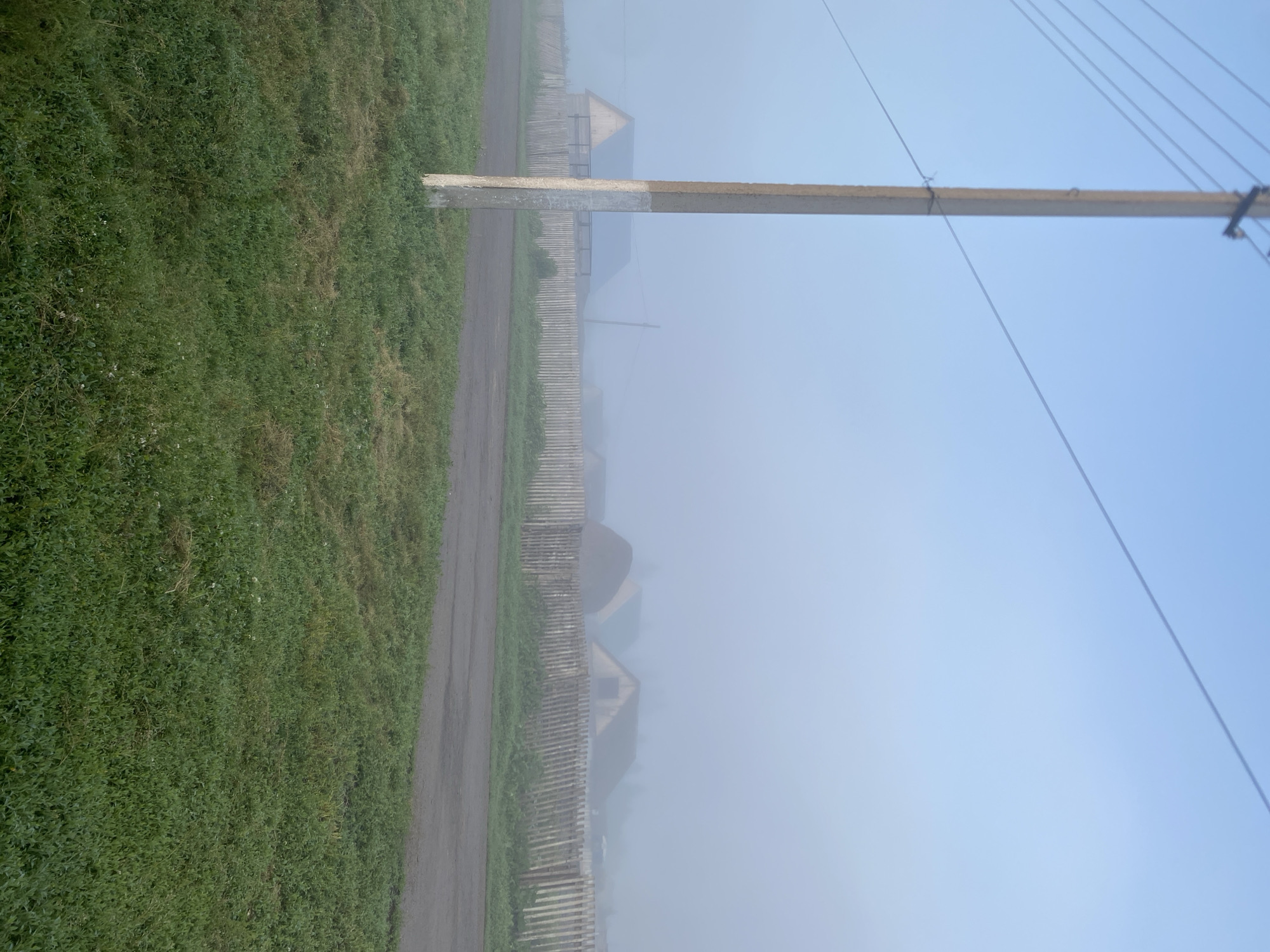A foggy morning in Maloarslangulovo, Russia, obscures a rural landscape. A concrete utility pole stands in the foreground, its lower section painted white. Beyond the pole, a paved road curves into the distance, bordered by green grass. A wooden fence runs parallel to the road, partially separating it from a cluster of houses shrouded in mist. The roofs of several structures are visible, their shapes softened by the fog. The sky above is a pale, hazy blue, suggesting the early hours of the day.