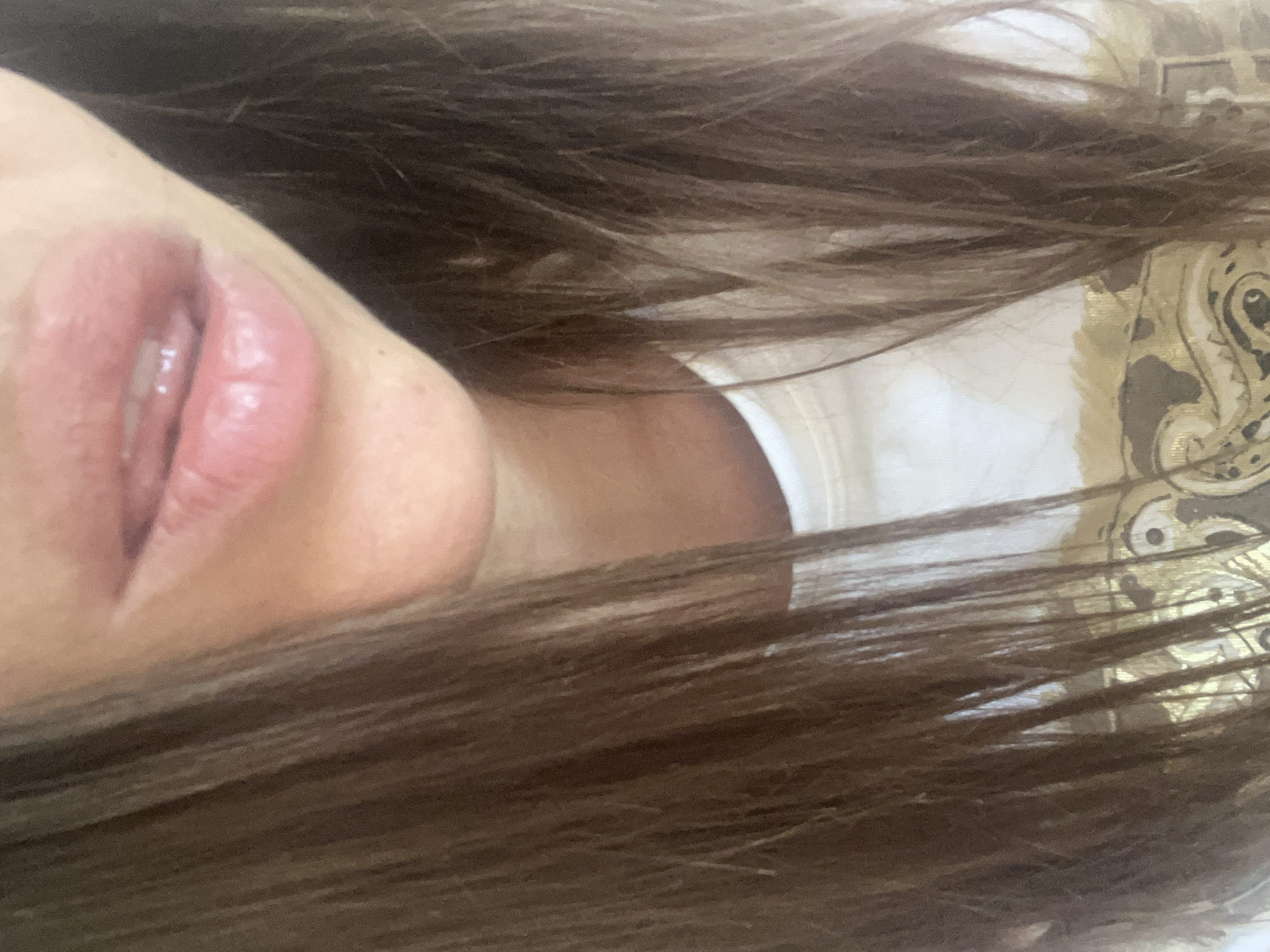 The image shows a close-up, upper-body view of a person with long, dark brown hair. The person is wearing a white t-shirt with a gold design. The focus is on the lower part of the face, specifically the lips, which appear to be slightly parted. The background is out of focus and appears to be an indoor setting. There is no indication of the location being in a city or any specific geographical area.