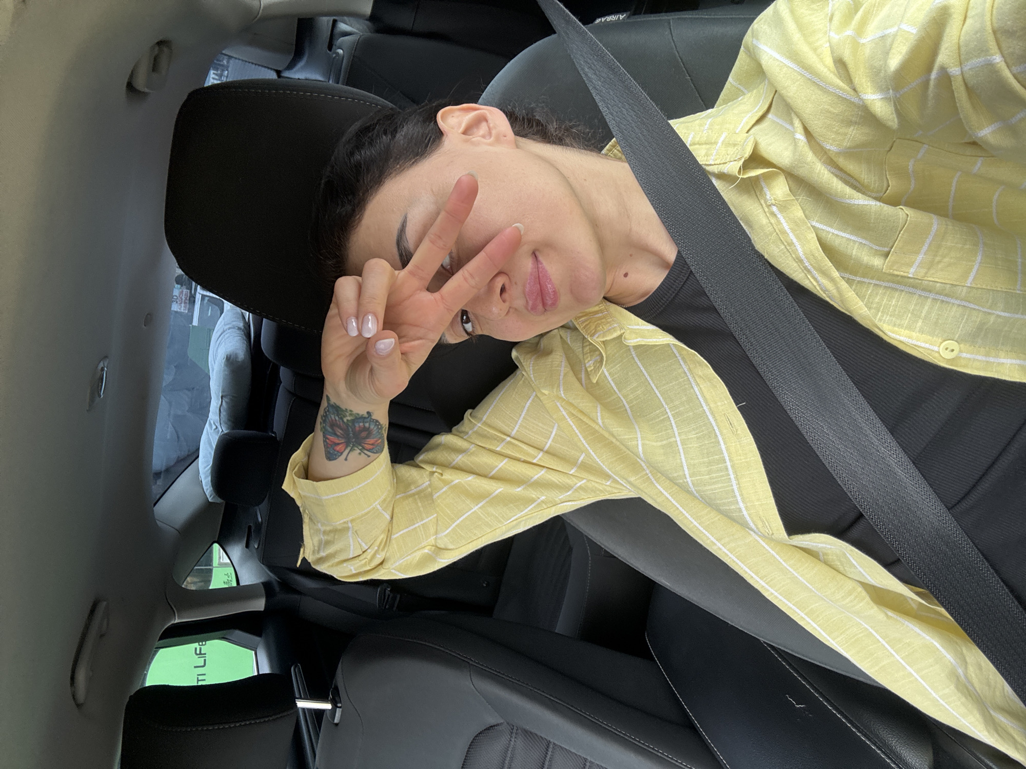 A young woman is captured in a selfie or photo, seated in the front passenger seat of a car. She has dark hair pulled back and is looking directly at the camera. Her left hand is raised, making a peace sign (V-sign) which partially obscures her face. She is wearing a yellow and white striped long-sleeved button-up shirt over a black top, with a seatbelt diagonally across her chest. A colorful butterfly tattoo is visible on her left forearm/wrist. Her nails are manicured in a light, possibly French style.
The setting is the interior of a modern car, featuring black leather seats, headrests, and a dark seatbelt. Through the car windows, blurred elements of an outdoor environment are visible, including distant buildings and some greenery, suggesting an urban or suburban area in Tbilisi, Georgia. The lighting indicates it is daytime.
Visible text includes "AIRBAG" on the car's pillar, and a partial green sign outside the car that appears to read "TI LIFE" or "ULTI LIFE."