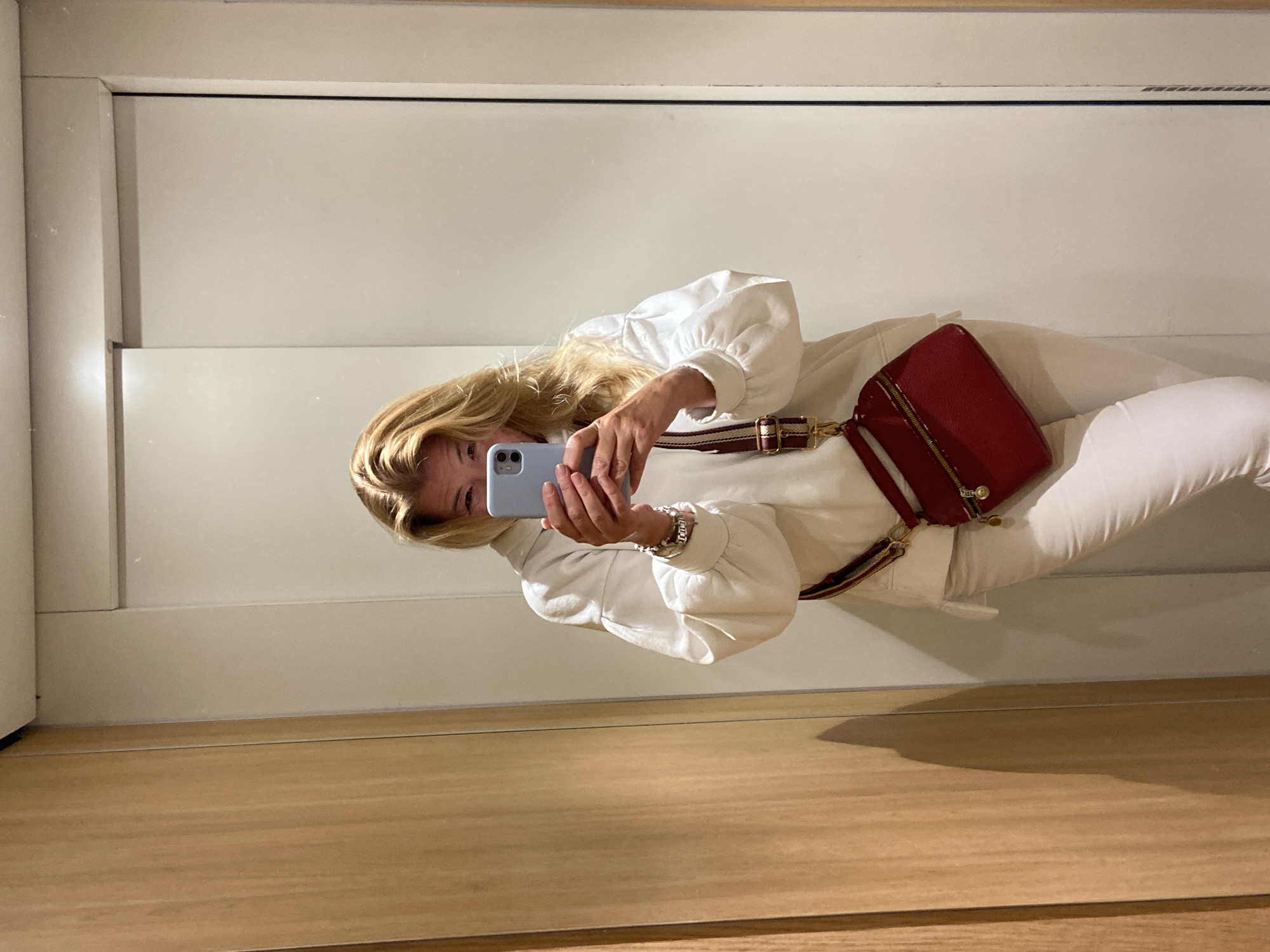 A woman is taking a picture of herself in a mirror. She is wearing a white jacket with puffy sleeves and white pants. She has a red purse with a striped strap slung across her body. She is holding a light blue smartphone in her hands, obscuring most of her face. The environment appears to be an interior space with a neutral color palette. The floor is wood, and the walls are white. The location is unknown.