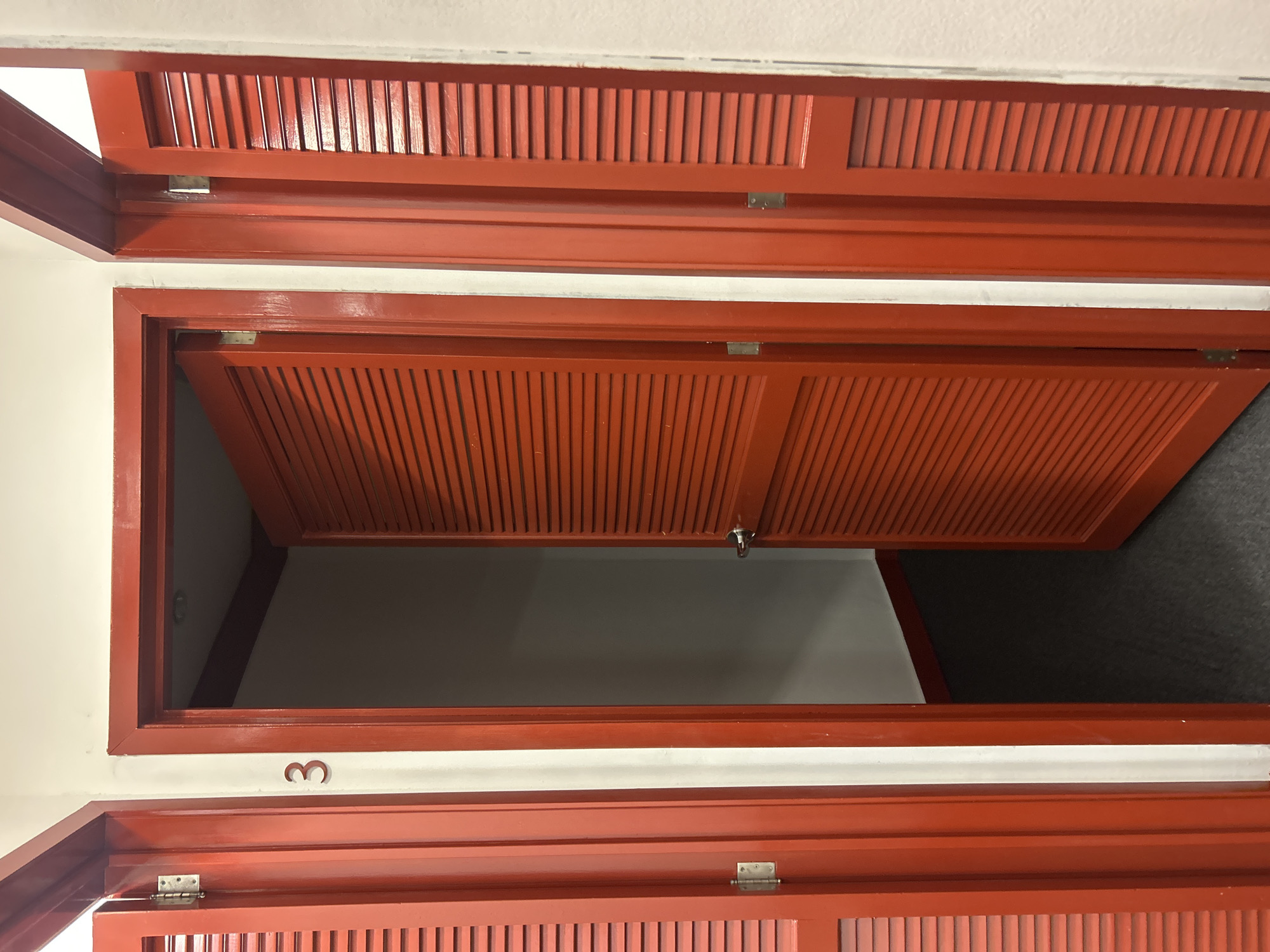 A low-angle interior shot depicts a series of red-painted wooden storage compartments or cabinets with louvered doors. One compartment is open, revealing a white interior and glimpses of what appears to be grey carpeting. The number "3" is marked on the wall underneath the open compartment. The location is unknown. There is no subject within this image to provide details about.