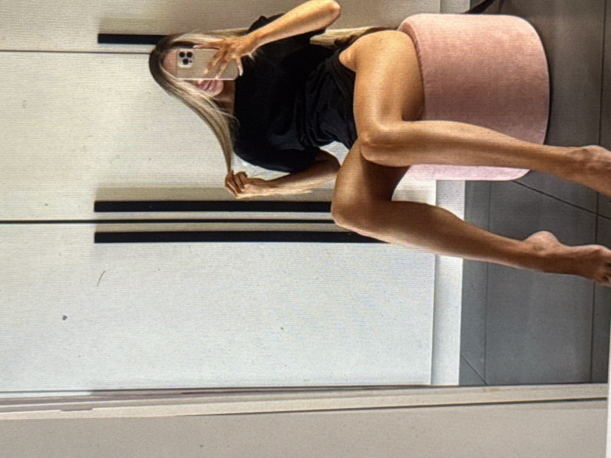 The image shows a woman taking a selfie in a room. She is sitting on a round, pink ottoman with a black t-shirt dress on. Her legs are extended, and she is holding a smartphone to take the picture. Her blonde hair is long and falls over her shoulder. The background of the image appears to be a closet with white doors and black handles. There are no specific location cues in the image to suggest Kuta Selatan, Indonesia. The lighting suggests it could be daytime indoors. The woman's expression is obscured by the phone, but she appears relaxed.