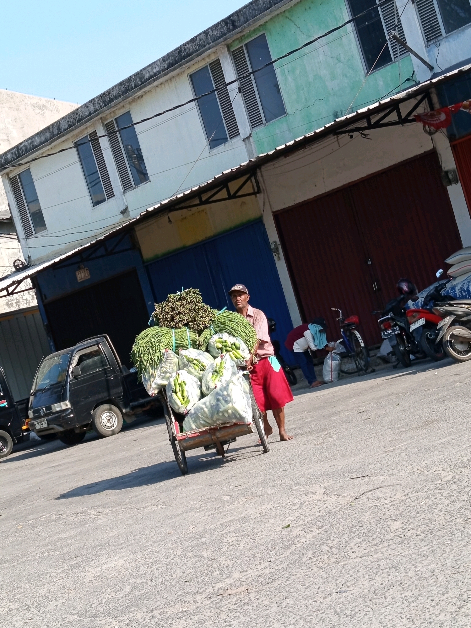 An outdoor, medium-shot photograph captures a street scene in Karawang, Indonesia, under a bright, clear sky. The foreground is dominated by a concrete road, with a shadow of a three-wheeled cart stretching across it. A man, wearing a light-colored shirt, a red skirt, and a baseball cap, stands to the right of the cart. He appears to be pushing or guiding the cart laden with a large quantity of vegetables, including green beans and possibly cucumbers.
Behind the man and the cart, a black pickup truck is parked. Further back, a row of buildings line the street. These buildings are two stories high and display a mix of architectural styles and colors. The building on the left is white, with several rectangular windows that have louvers. The building next to it is painted a pale green and also features similar windows. The facade of these buildings shows some signs of age and weathering.
In front of the buildings, a covered walkway with a sloped roof extends along the storefronts. The storefronts themselves appear to be dark, possibly garage doors or large entrances. Several motorcycles are parked in front of one of the buildings on the right side of the image, one of which is red. Another individual, wearing a maroon and white top with a blue scarf or cloth, is visible bending down near a bicycle, seemingly interacting with a plastic bag. The overall impression is that of a market area or a commercial street in a town.