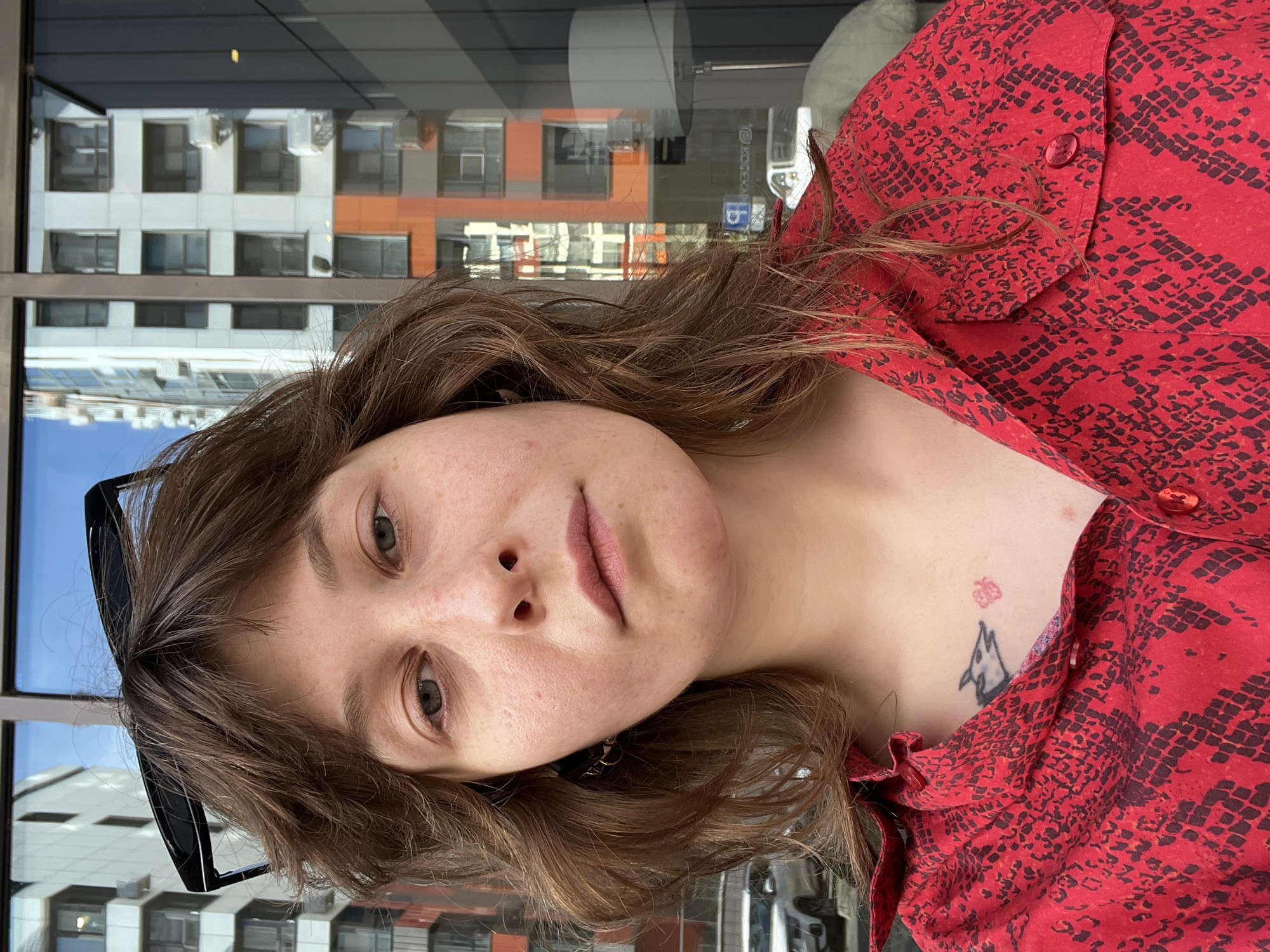 A medium-shot, head-and-shoulders selfie captures a woman with brown, shoulder-length hair and sunglasses perched on her head. She is wearing a red button-up shirt with a dark, reptilian pattern. A small tattoo of a fox and a red flower is visible on her upper chest. The woman is positioned in front of a glass building with reflections of other buildings and a street with cars. The scene appears to be outdoors in a city environment.