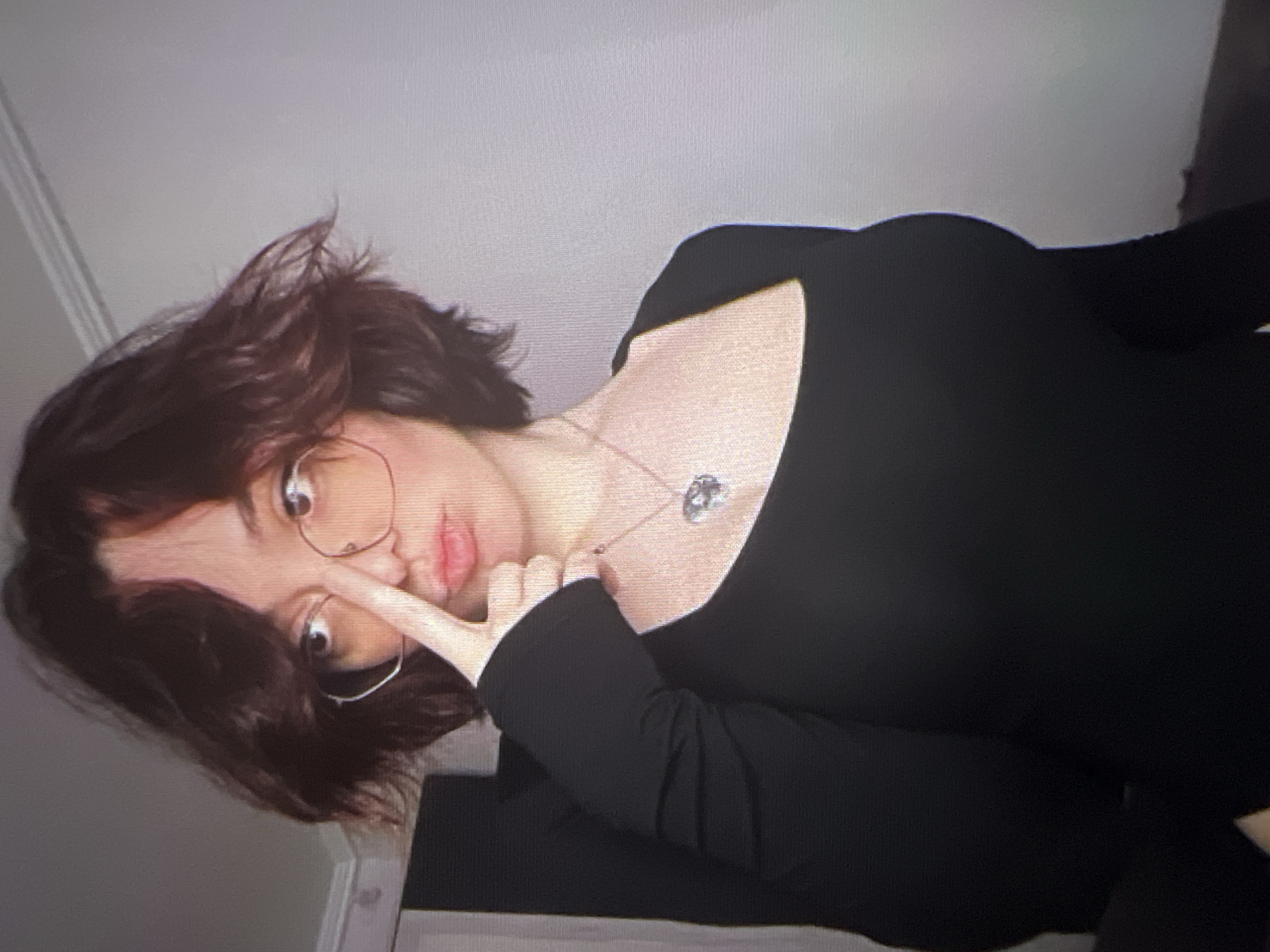 A young woman with shoulder-length, dark brown, messy hair and bangs is captured in a close-up shot. She is wearing round, light-colored glasses and a black, long-sleeved, square-neck top with a silver heart-shaped pendant necklace. Her right hand is raised, with her index finger touching the bridge of her nose. Her lips are slightly parted. The background is a plain, light-colored wall, suggesting an indoor setting. There is no identifiable information about the location in Energetik, Russia.