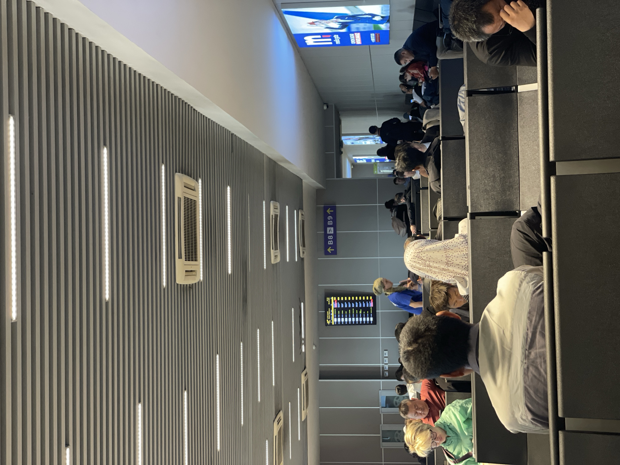 The image depicts an airport waiting area, likely in Tashkent, Uzbekistan. Passengers are seated in rows of black chairs. A wall with vertical gray panels interspersed with vertical LED light strips runs along the left side of the image, punctuated by air conditioning units. Overhead signage directs passengers toward gates B8 and B9. A flight information display board is visible. The area appears modern and well-lit.