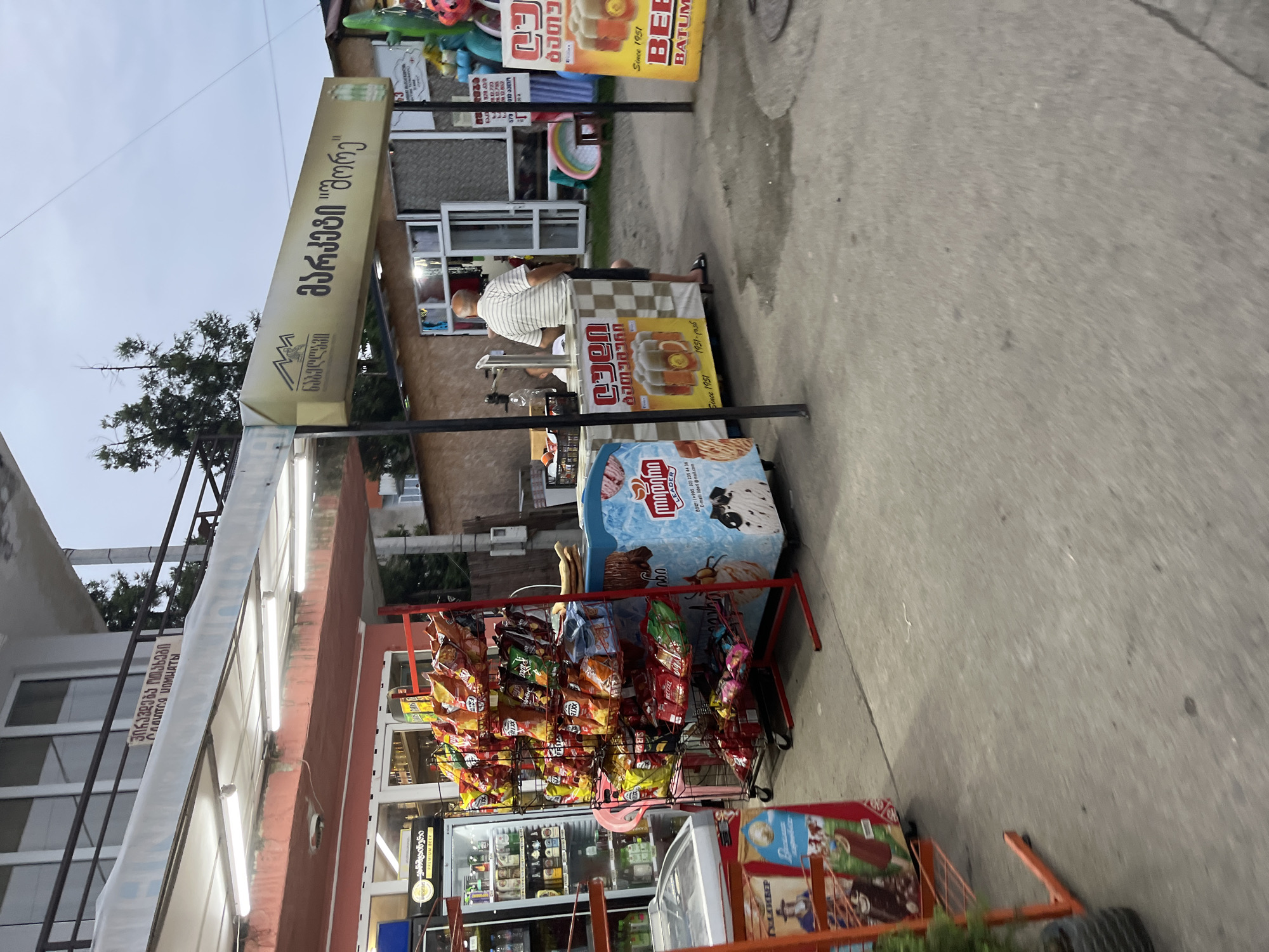 This is a street-level, medium-shot photograph taken outdoors in Ureki, Georgia, likely in the evening given the artificial lighting. The scene depicts a small, open-air market or convenience store.
In the foreground, to the left, is an orange metal rack filled with rows of colorful snack bags. To the right of the snack rack is a blue, refrigerated display case advertising ice cream, with images of cones and a cow. Further to the right, there's a table with a checkered cloth, behind which a man wearing a striped shirt and shorts is standing. He appears to be an employee or vendor. The table has a banner displaying an image of beer steins and text in Georgian. Another similar banner for beer is visible further to the right, mounted on a pole.
In the background, a building with multiple windows and a balcony is visible. A large, illuminated sign with Georgian text and a mountain logo hangs from the awning of the market. The ground is paved with concrete. The overall impression is a typical street-side commercial area in a resort town, catering to tourists.