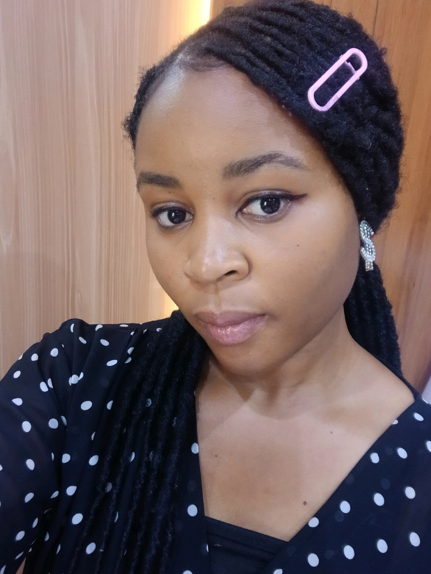 The image shows a close-up, head-and-shoulders portrait of a young Black woman. She has dreadlocks styled back from her face, secured with a pink barrette on the right side. She is wearing a black dress with white polka dots and a sparkling earring in the shape of a dollar sign on her left ear. Her gaze is directed towards the viewer. The background consists of a wooden wall. The lighting suggests an indoor setting. The image is taken from a slightly upward angle.