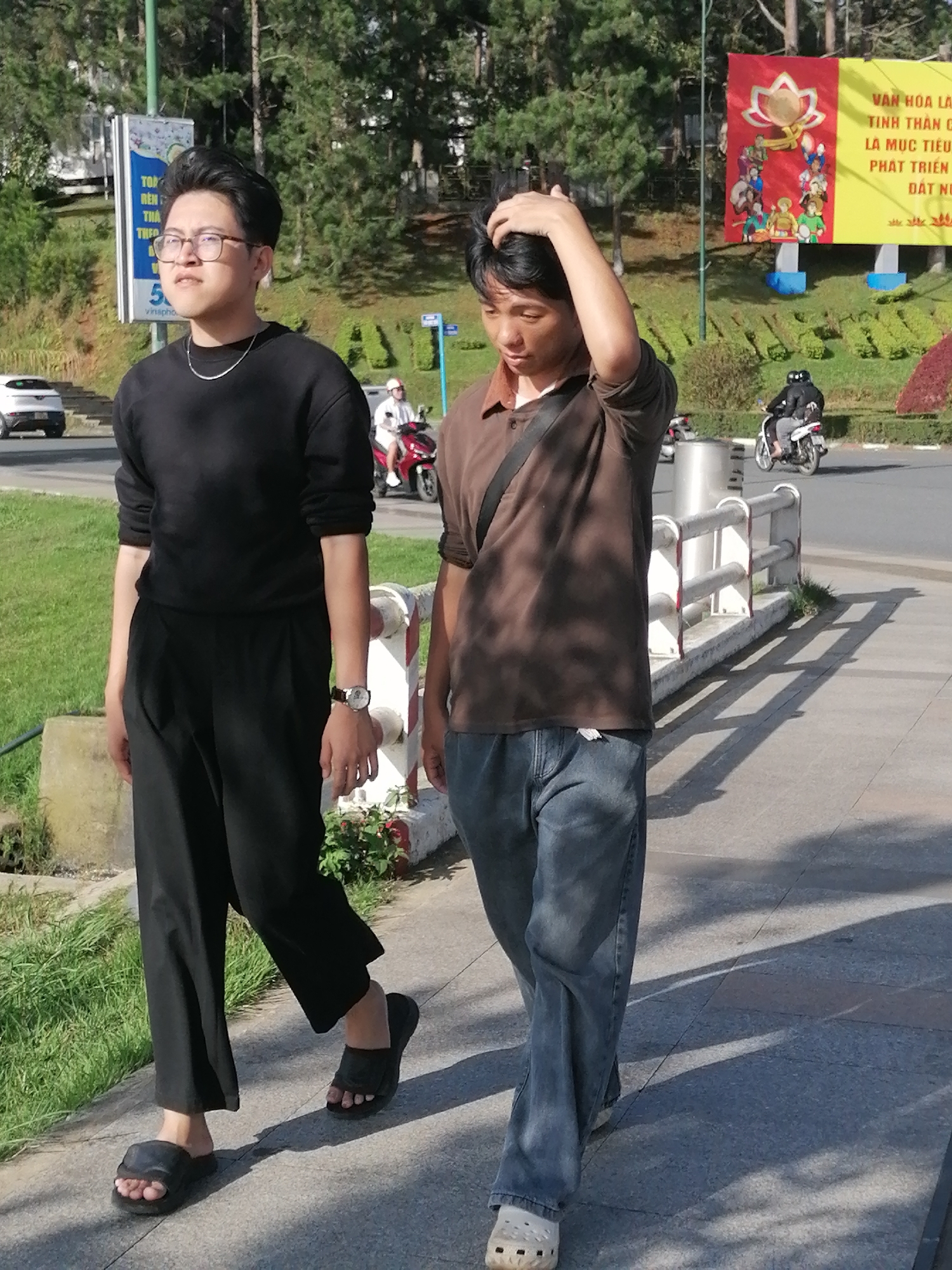 Two young men walk along a paved sidewalk in Da Lat, Vietnam. The man on the left, wearing glasses, a black long-sleeved shirt, and black pants, walks in black sandals. He has a silver necklace on. The man on the right, in a brown short-sleeved polo shirt and blue jeans, walks in white clogs. He is touching his head with his right hand. Behind them, a busy street with a white car and a red motorcycle can be seen. A billboard with Vietnamese writing and images of people and a lotus flower is visible in the background. The environment is sunny with shadows cast on the ground by trees and a railing.