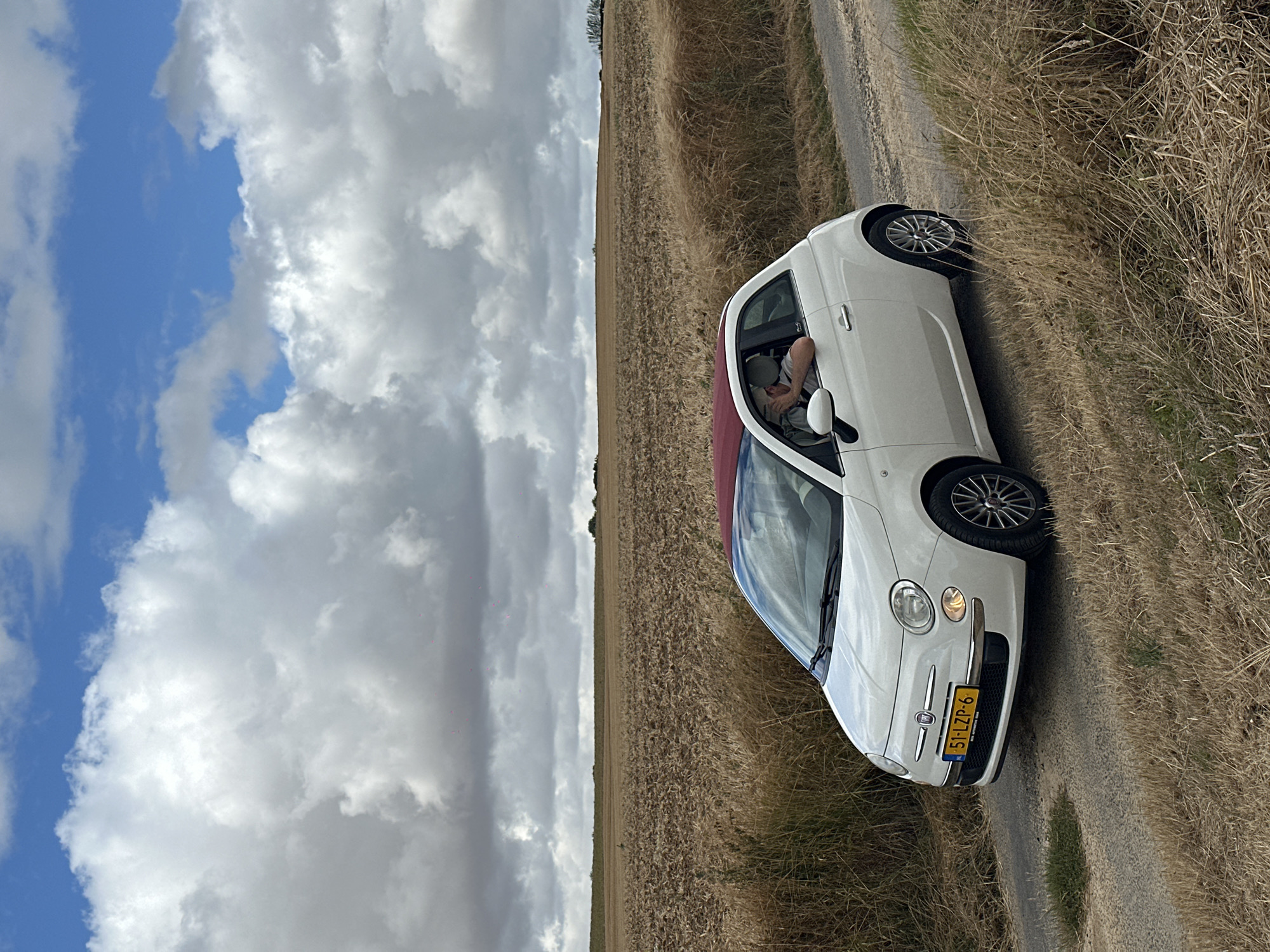 In Wambercourt, France, on a cloudy day, a white Fiat 500 convertible with a red roof is parked on a dirt road next to a dry, grassy field. The car's license plate reads "51-LZP-6". A person wearing a light-colored shirt and a cap is leaning out of the driver's side window, appearing to adjust their hat. The background shows a harvested agricultural field and a distant treeline under a dramatic sky filled with fluffy white and grey clouds.