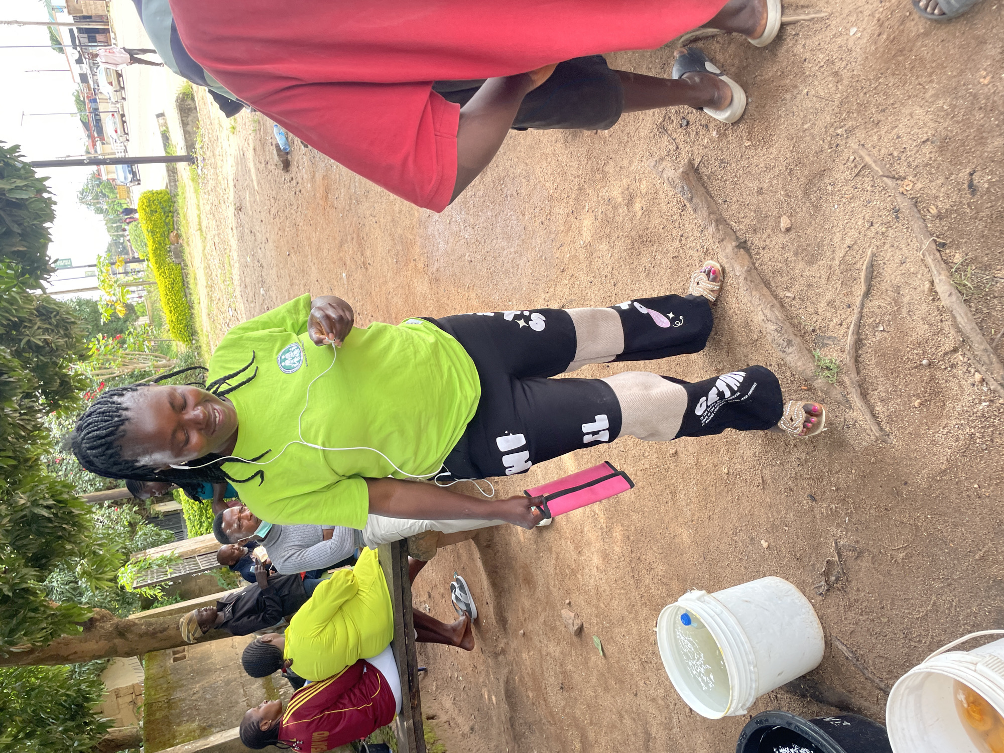 A group of people are gathered outdoors in what appears to be a semi-rural area of Jos, Nigeria. The scene is unpaved with scattered debris and vegetation. In the foreground, a woman wearing a bright green t-shirt and black patterned pants is holding a pink object and listening to music through earbuds. Behind her, a person in a red shirt is partially visible. Several other individuals are seated on a wooden bench in the middle ground. One woman wears a maroon and yellow tracksuit, and another is in a vibrant yellow garment. A man in a gray sweater is also seated on the bench. In the background, a street with vehicles and buildings suggests proximity to a more urban area. Two buckets, one filled with water, are visible on the ground in the foreground, indicating a possible water distribution or collection point. The overall impression is a candid snapshot of daily life in Jos.
